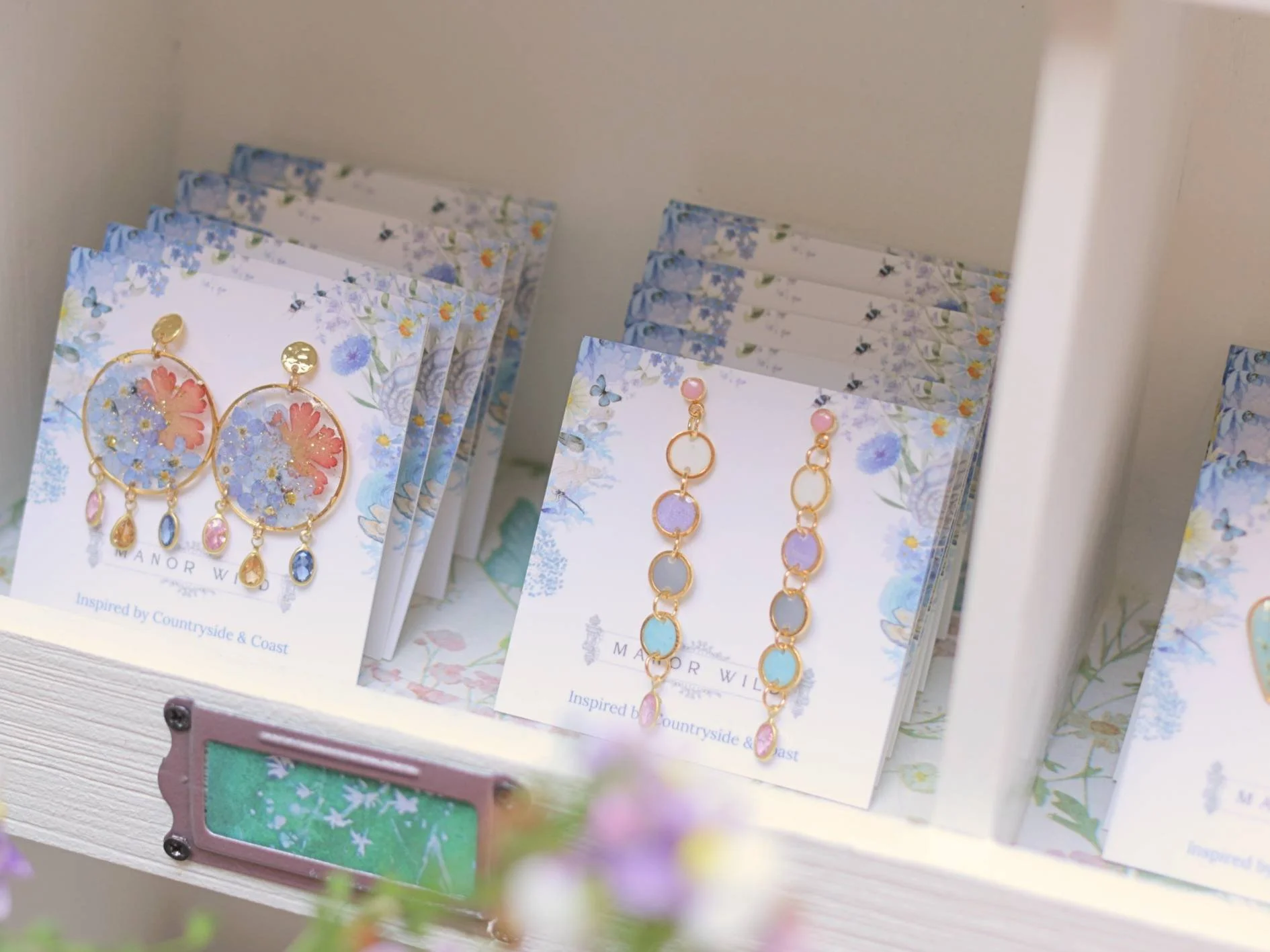 Display of colorful earrings with floral and butterfly designs on paper inserts, arranged on a white shelf or display stand, with some blurred plants in the foreground.