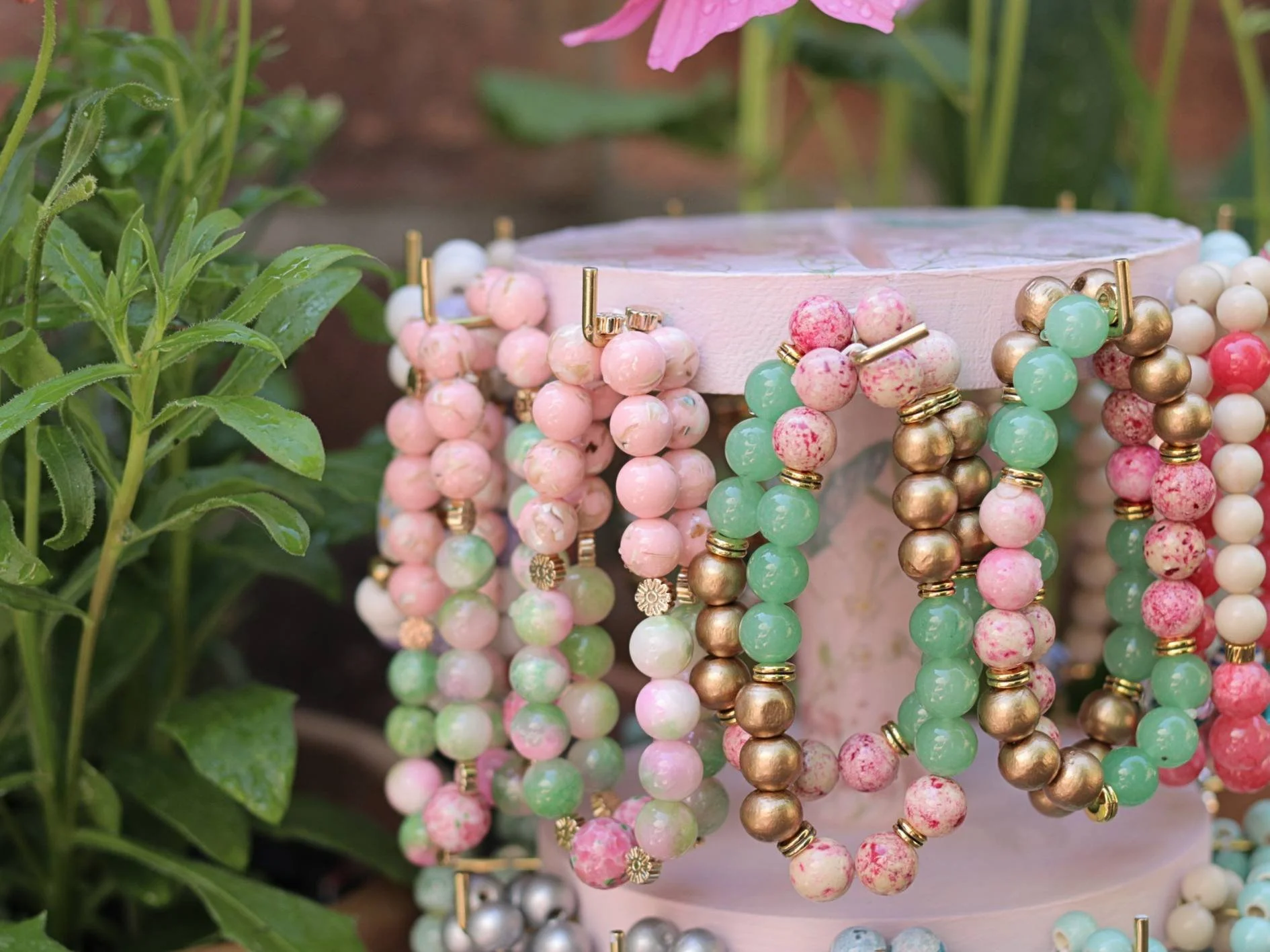 Display of pastel-colored beaded bracelets hanging on a stand, surrounded by green foliage, with a pink flower above.
