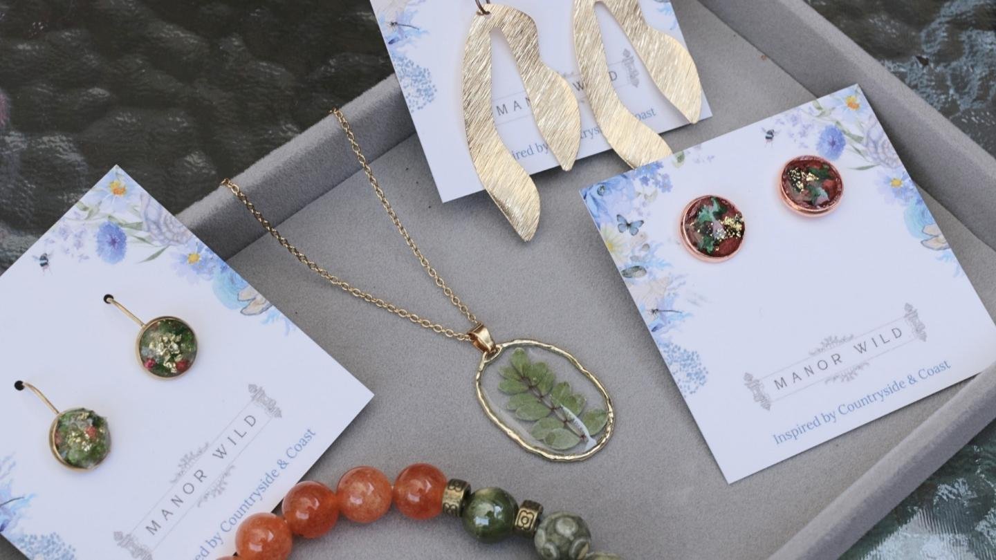 Jewelry display featuring earrings, necklaces, and bracelets with green and floral details on cards labeled 'Manor Wild' and inspired by countryside and coast.