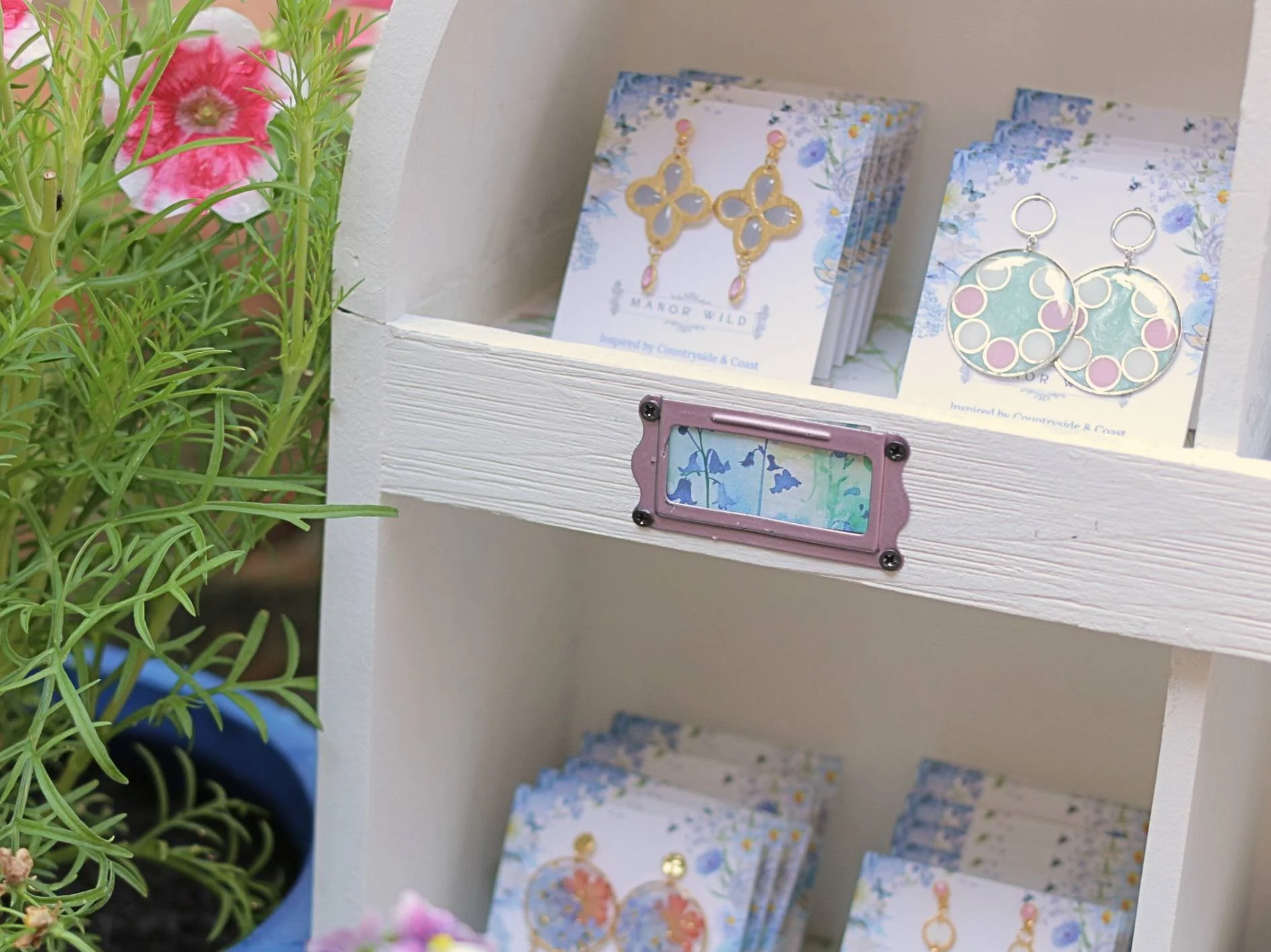 Display shelf with assorted jewelry and greeting cards, next to a potted green plant with pink and white flowers.