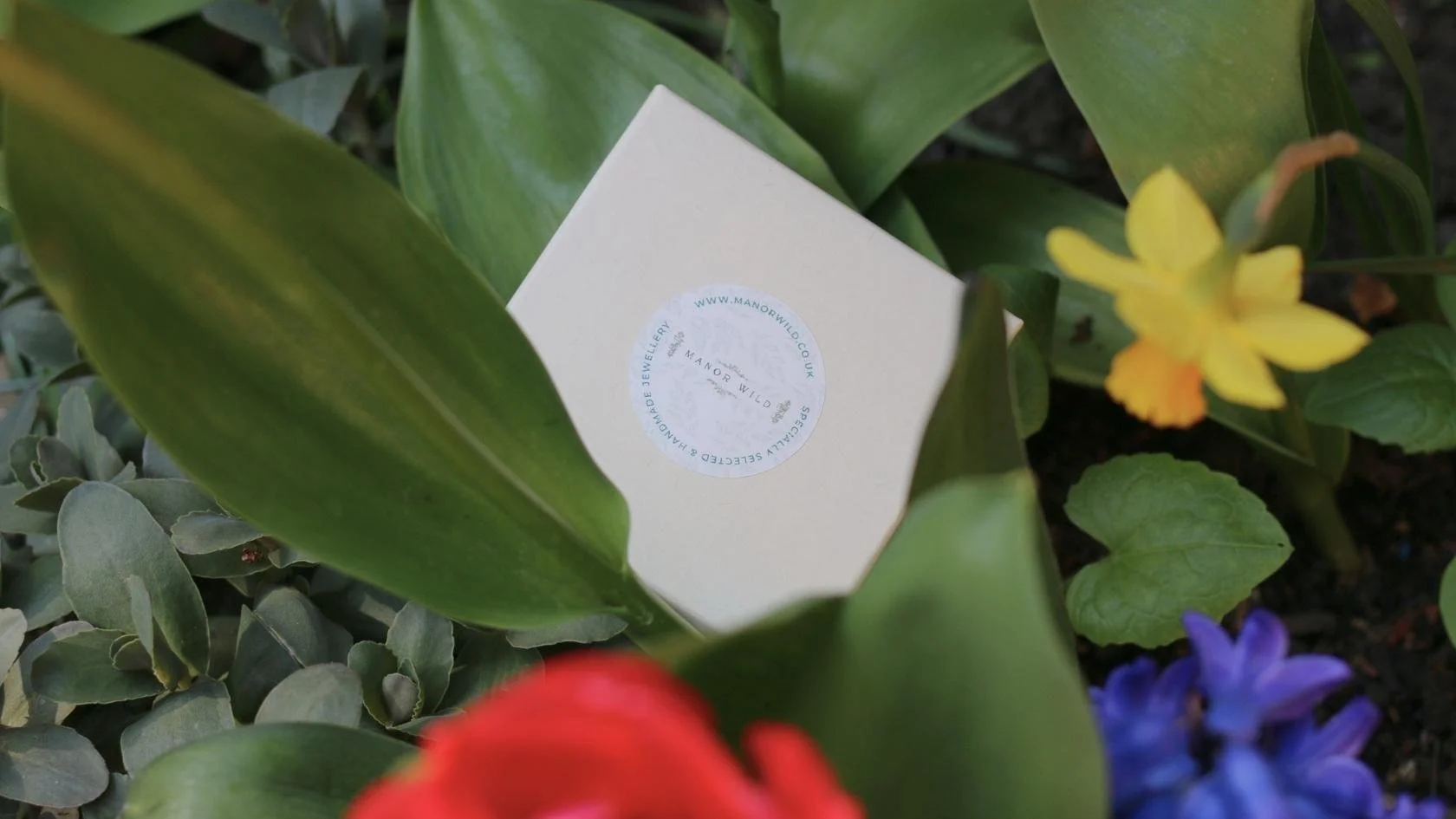 A white box with a circular sticker on top amidst green leaves and colorful flowers.