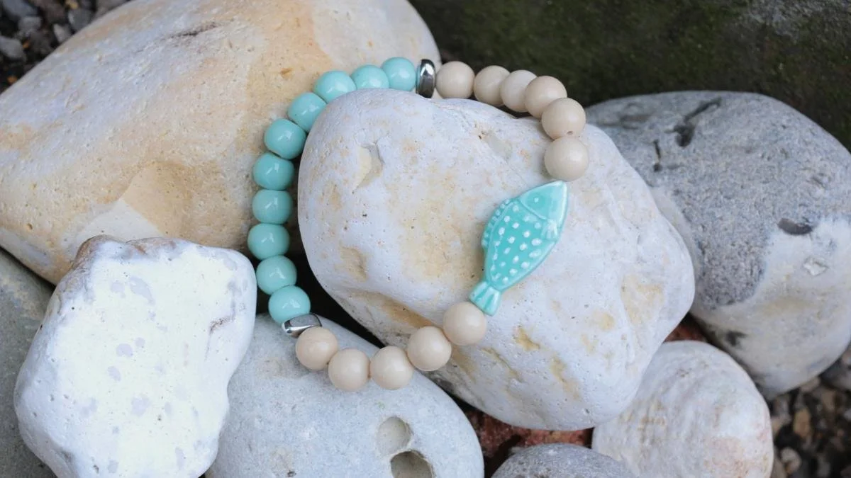 A beaded bracelet with a light blue fish charm, resting on white and beige rocks outdoors.