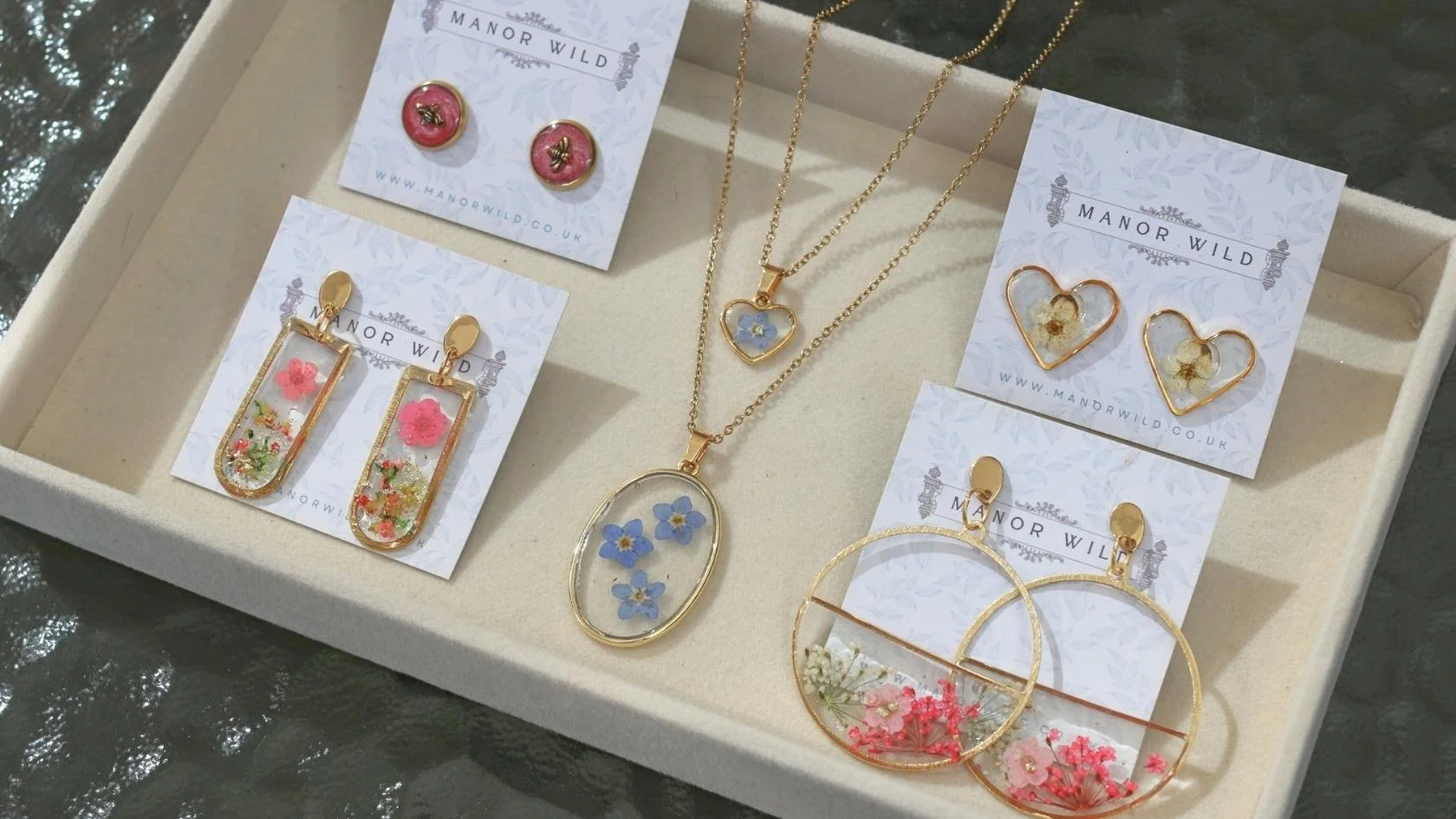 Jewelry box containing assorted gold earrings and necklaces with pressed flowers inside the jewelry, labeled with 'Manor Wild' branding.
