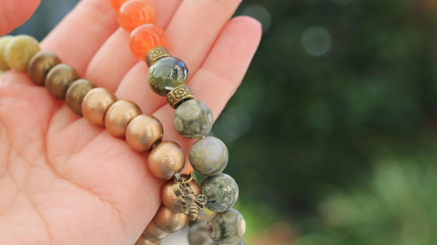 Close-up of a hand holding a beaded bracelet with green, brown, and orange beads, against a blurred green background.