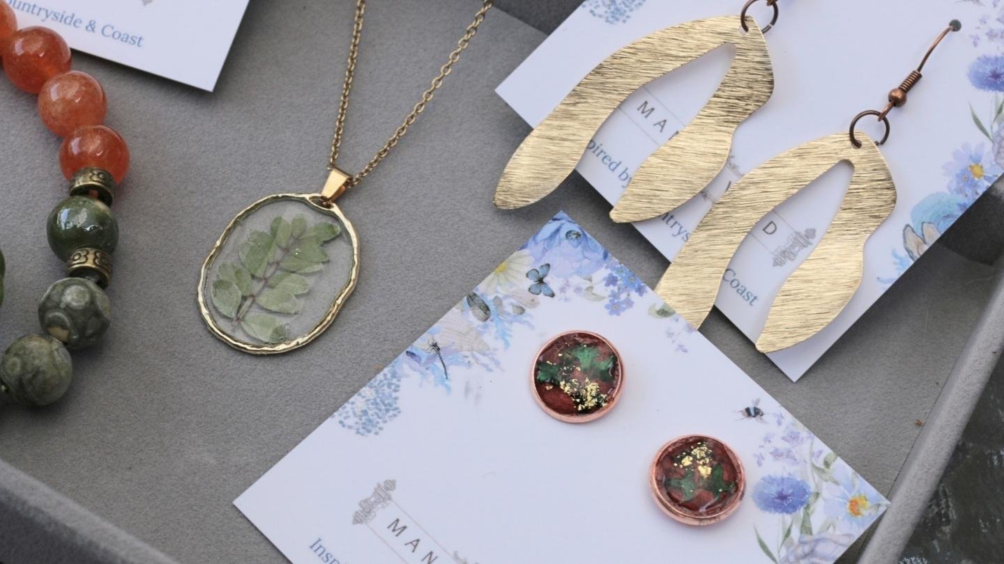 Assorted jewelry including a beaded bracelet, a gold necklace with a green leaf pendant, gold leaf-shaped earrings, and circular earrings with a green and gold design, all displayed on a surface with floral cards.