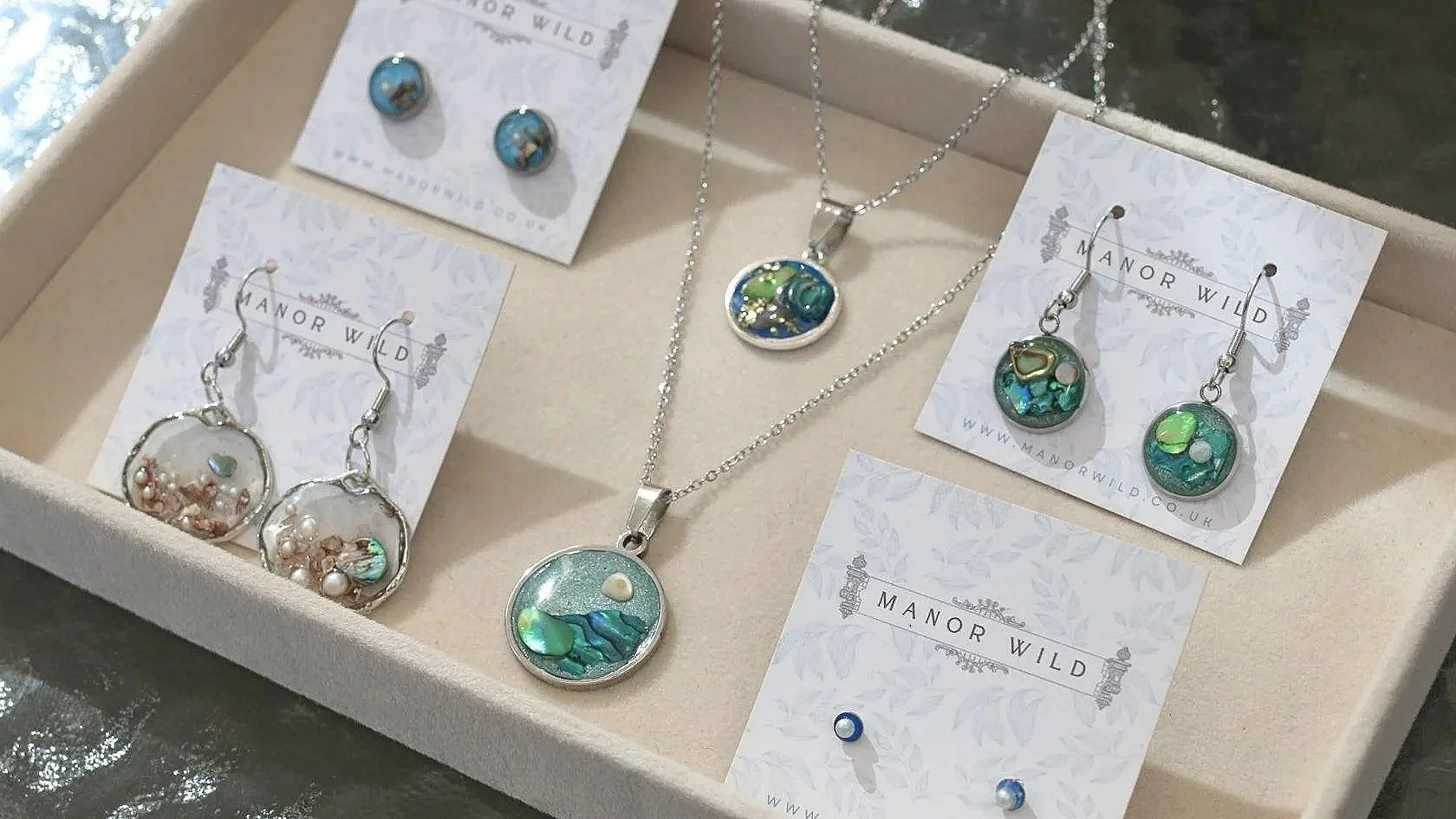 A jewelry set displayed in a beige gift box, including earrings, necklaces, and pendants with blue, green, and pearl accents from Manor Wild.