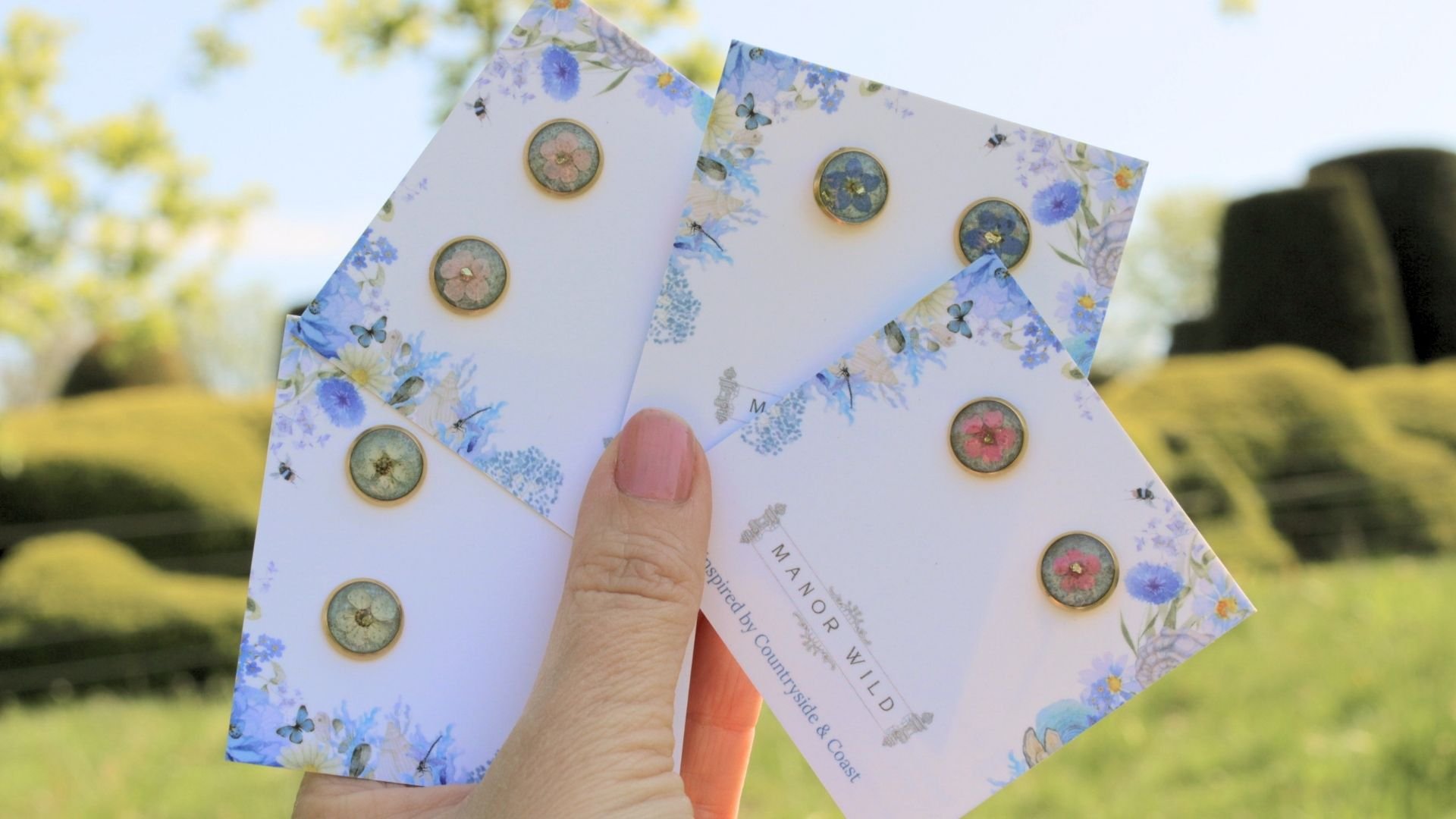 Person holding floral note cards with pressed flowers and bee illustrations outdoors on a sunny day.
