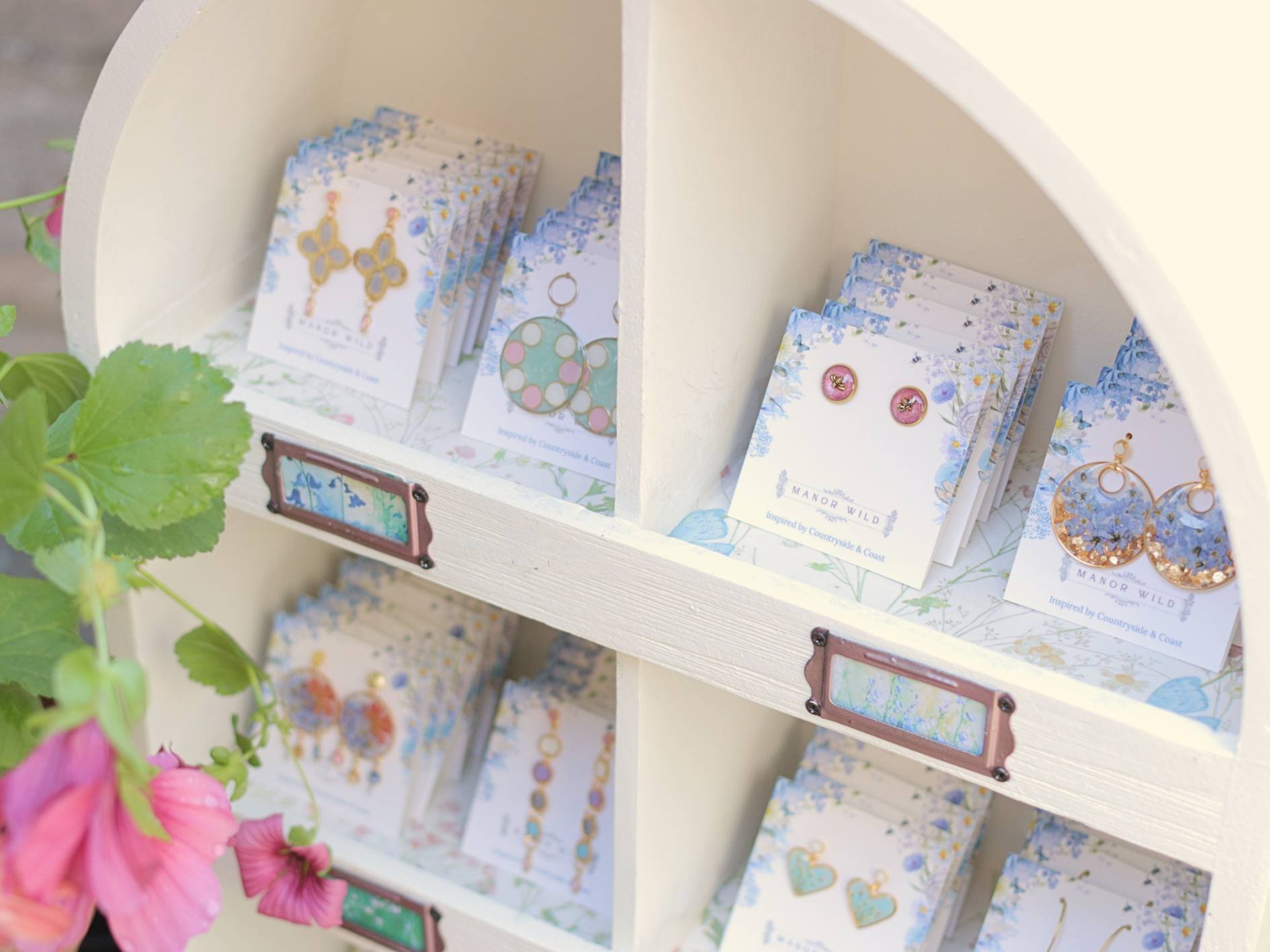 Display of jewelry cards on a white wooden shelf with pink flowers in the foreground.