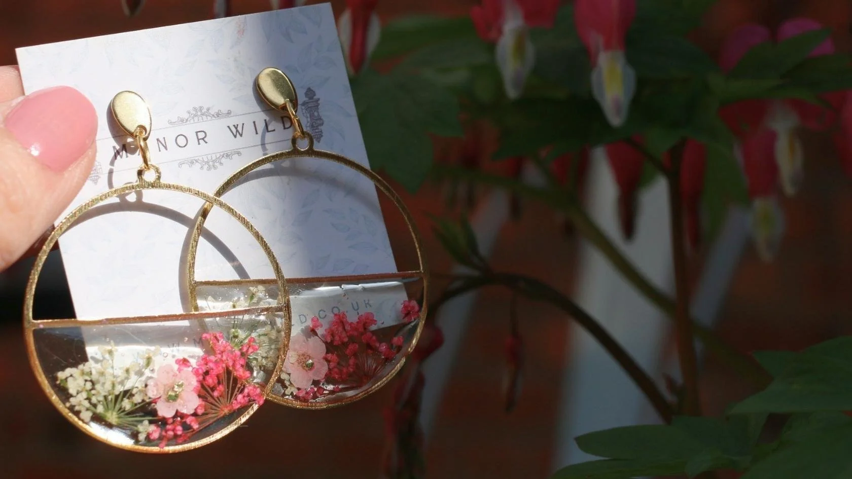 Gold hoop earrings with floral accents inside, attached to a display card with text, held up against a background of green leaves and red flowers.