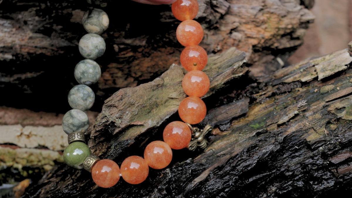 A beaded bracelet with orange, gray, and green marble-like beads and a small metallic flower charm, resting on a piece of bark.