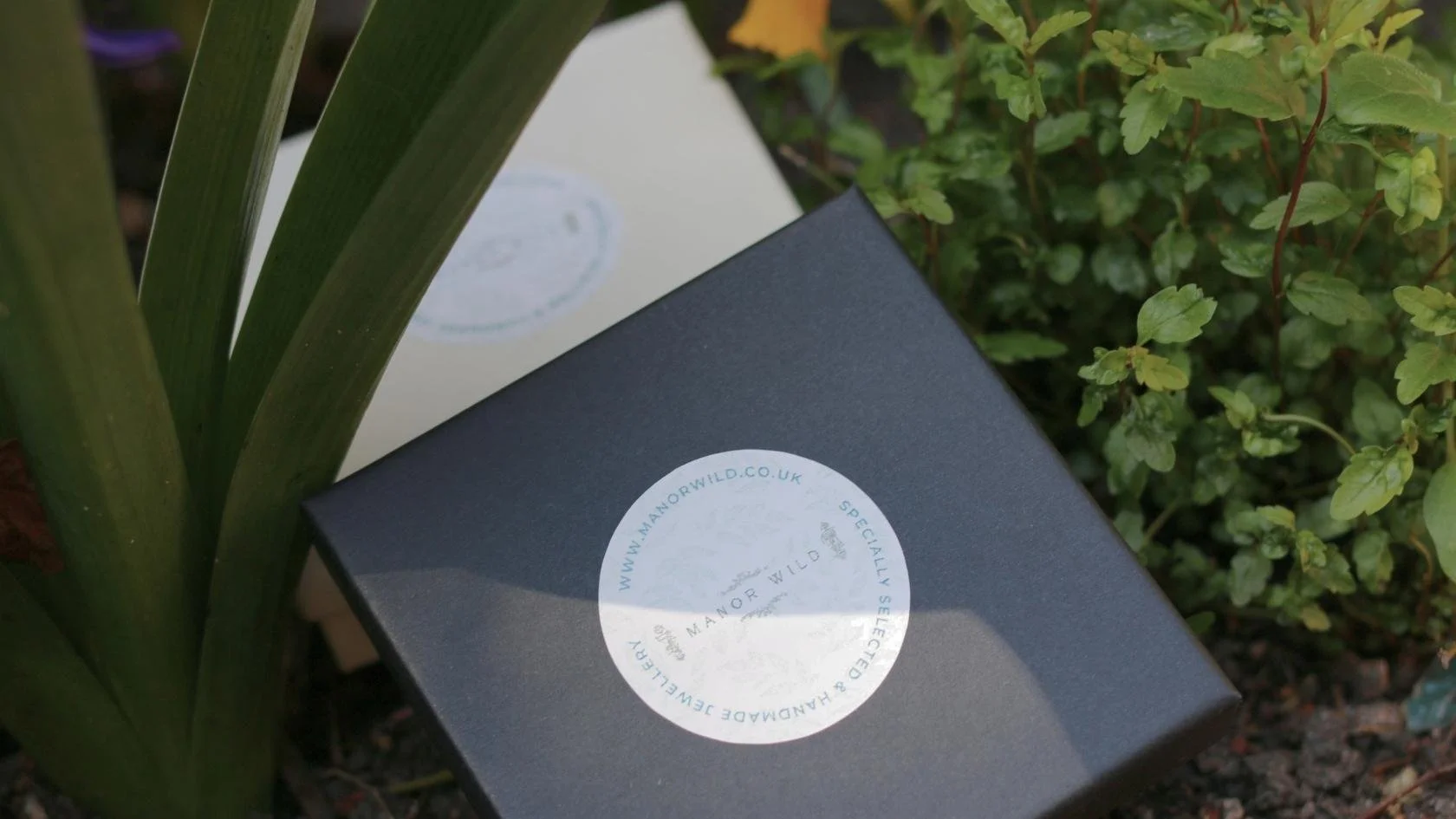 A dark gray box with a circular white sticker from Manor Wild, placed among green plants and flowers in a garden.