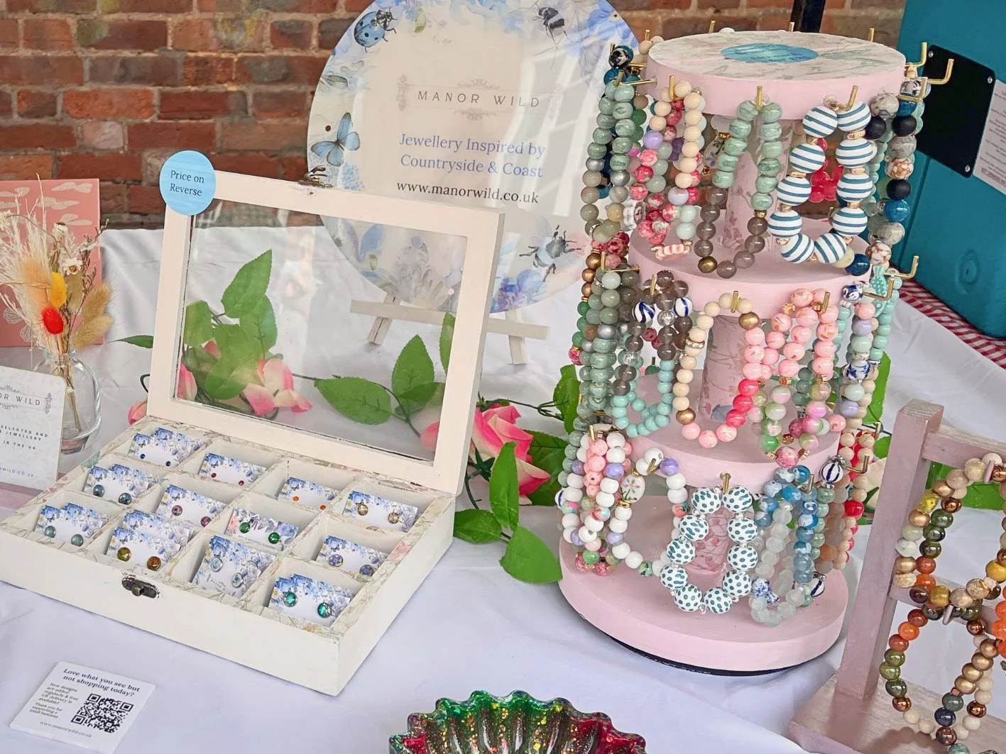 A display of colorful beaded jewelry on stands and trays at a craft market, with a brick wall in the background and a sign reading 'Manor Wild' behind the jewelry.