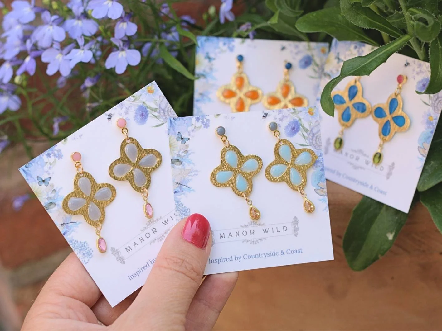 Pair of gold earrings shaped like butterflies with pastel colored stones, held by a hand with painted red nails, against backdrop of purple flowers.