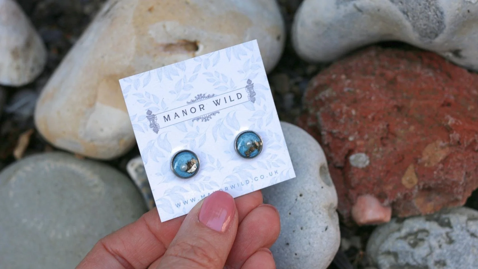 Pair of blue and gold round earrings on a white card marked 'Manor Wild' against rocks.