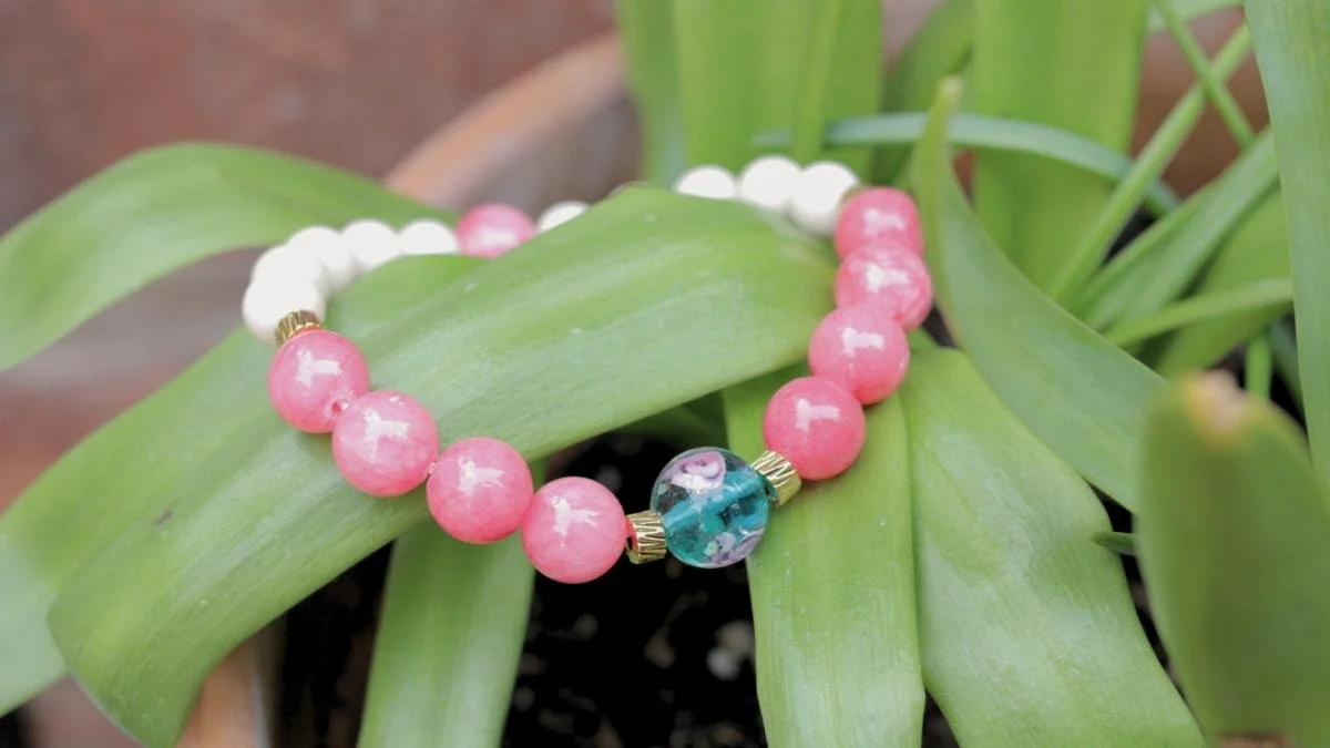 A beaded bracelet with pink, white, and a multicolored blue-green bead resting on green leaves of a potted plant.