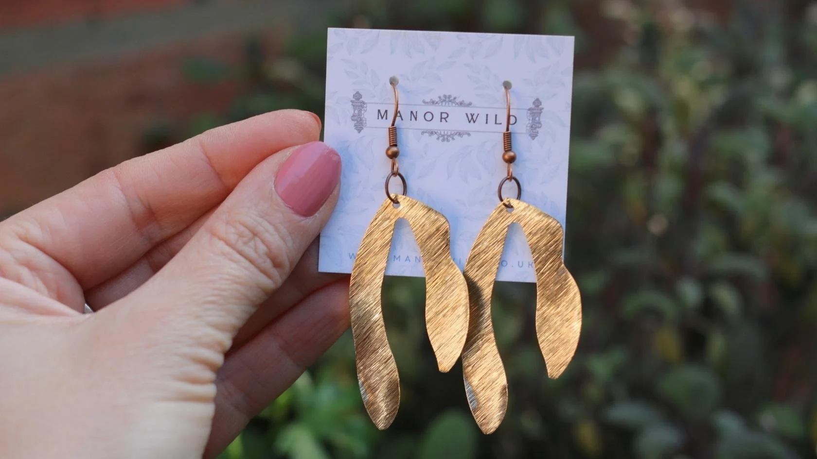 Gold-colored feather-shaped earrings on a white card labeled 'Manor WILD', held by a person with pink nails, outdoor background.