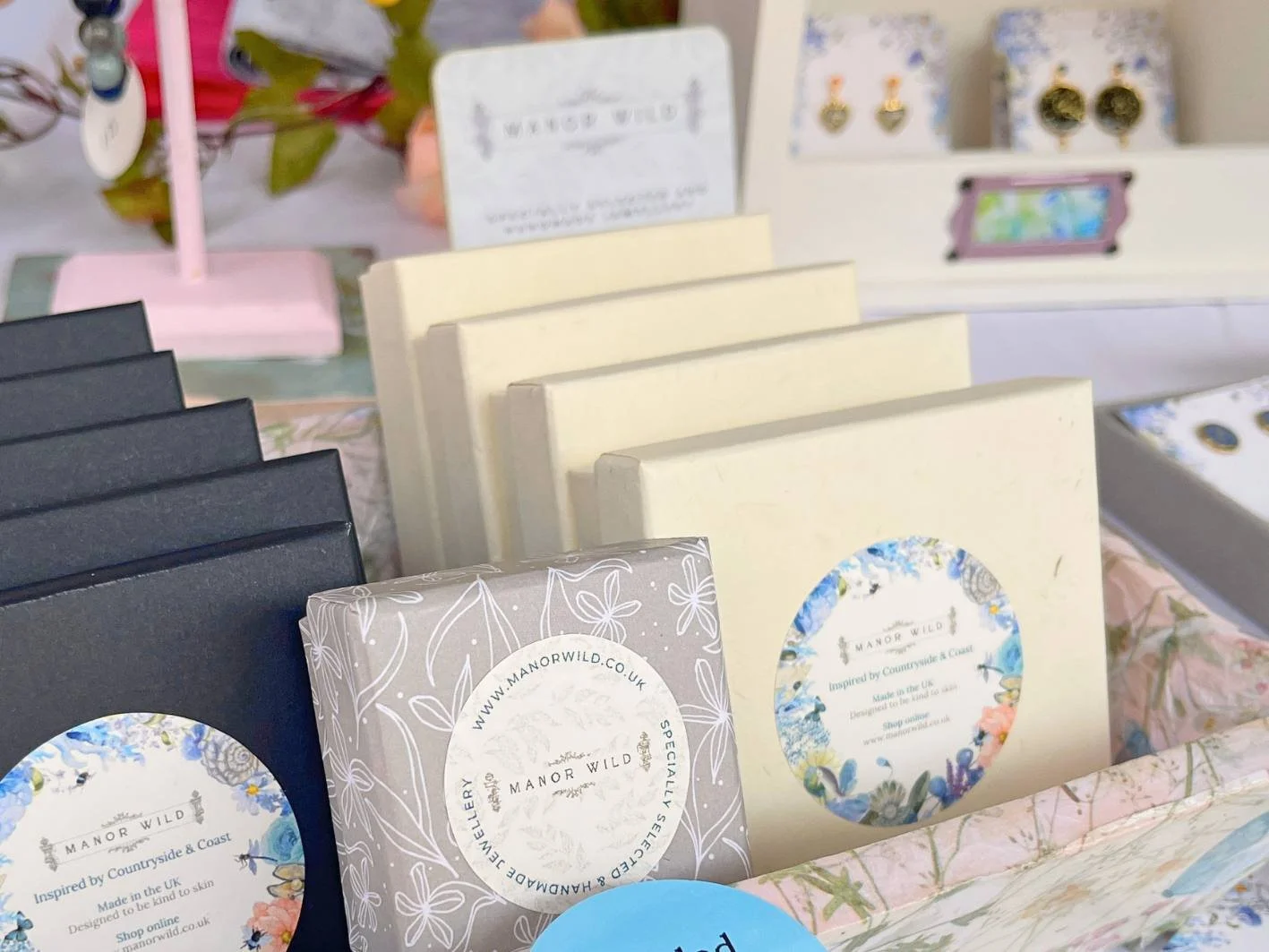 Display of Manor Wild jewelry and accessories in packaging, with some black and cream boxes, and a floral-patterned tray, in a retail setting.
