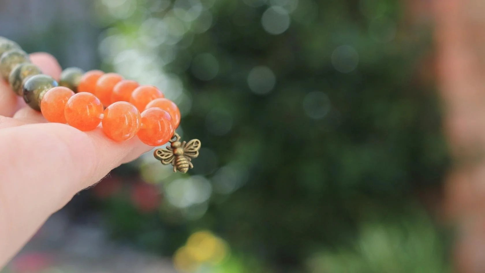 Close-up of a beaded bracelet with orange and green beads and a small brass bee charm, held by a person's hand against a blurred outdoor background.