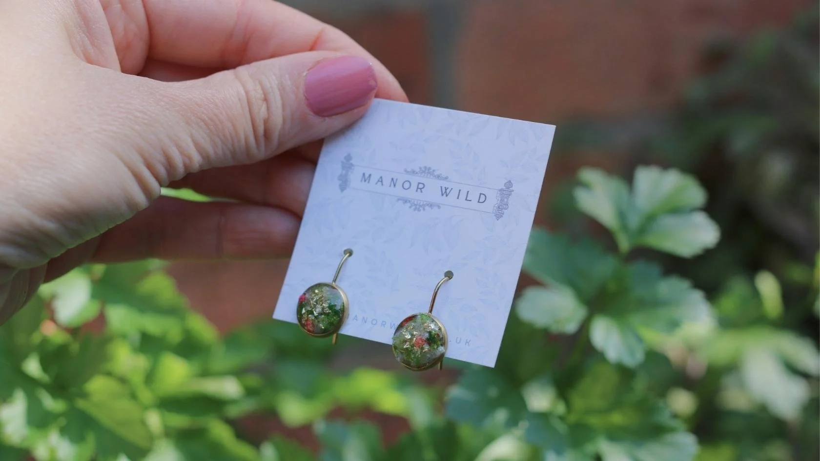 Hand holding a card with a pair of floral earrings displayed against green leafy background.