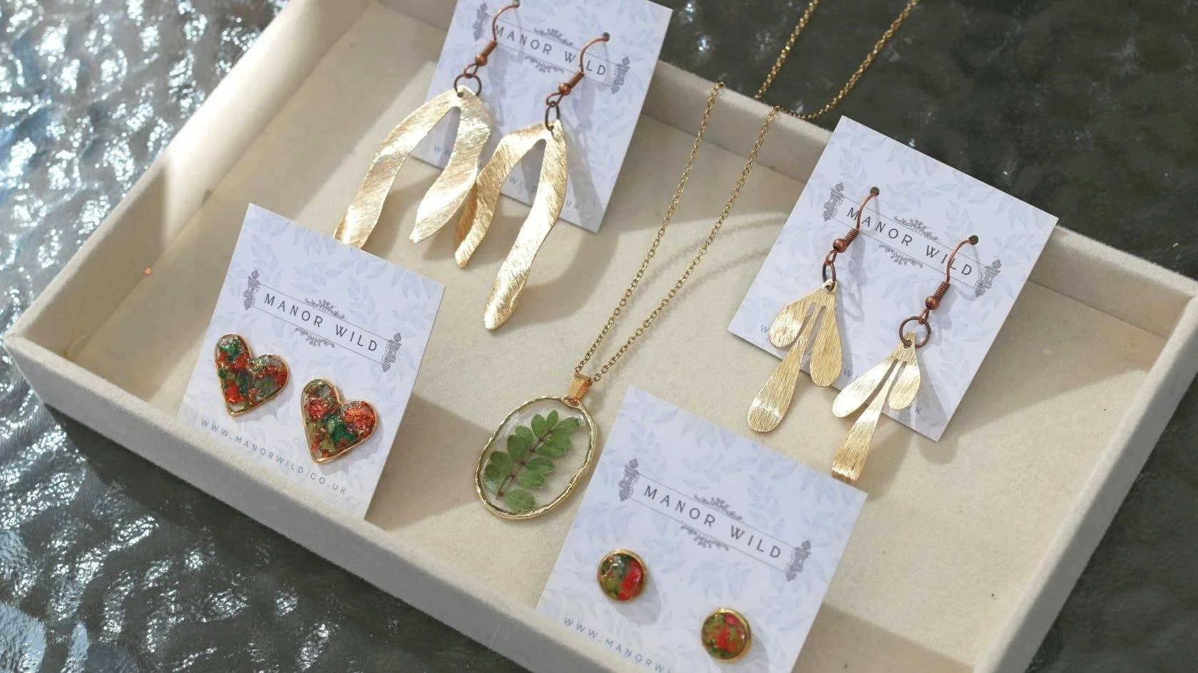 Jewelry display with gold leaf earrings, a pendant necklace with green leaves in resin, and matching leaf and heart earrings on cards labeled 'Manor Wild' inside a jewelry box.