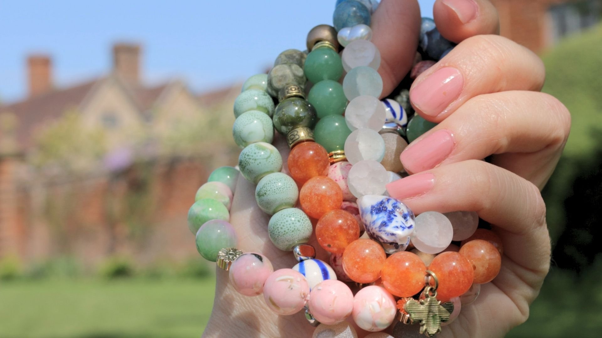 Close-up of a hand holding multiple colorful beaded bracelets outdoors with a blurry background of houses and trees.