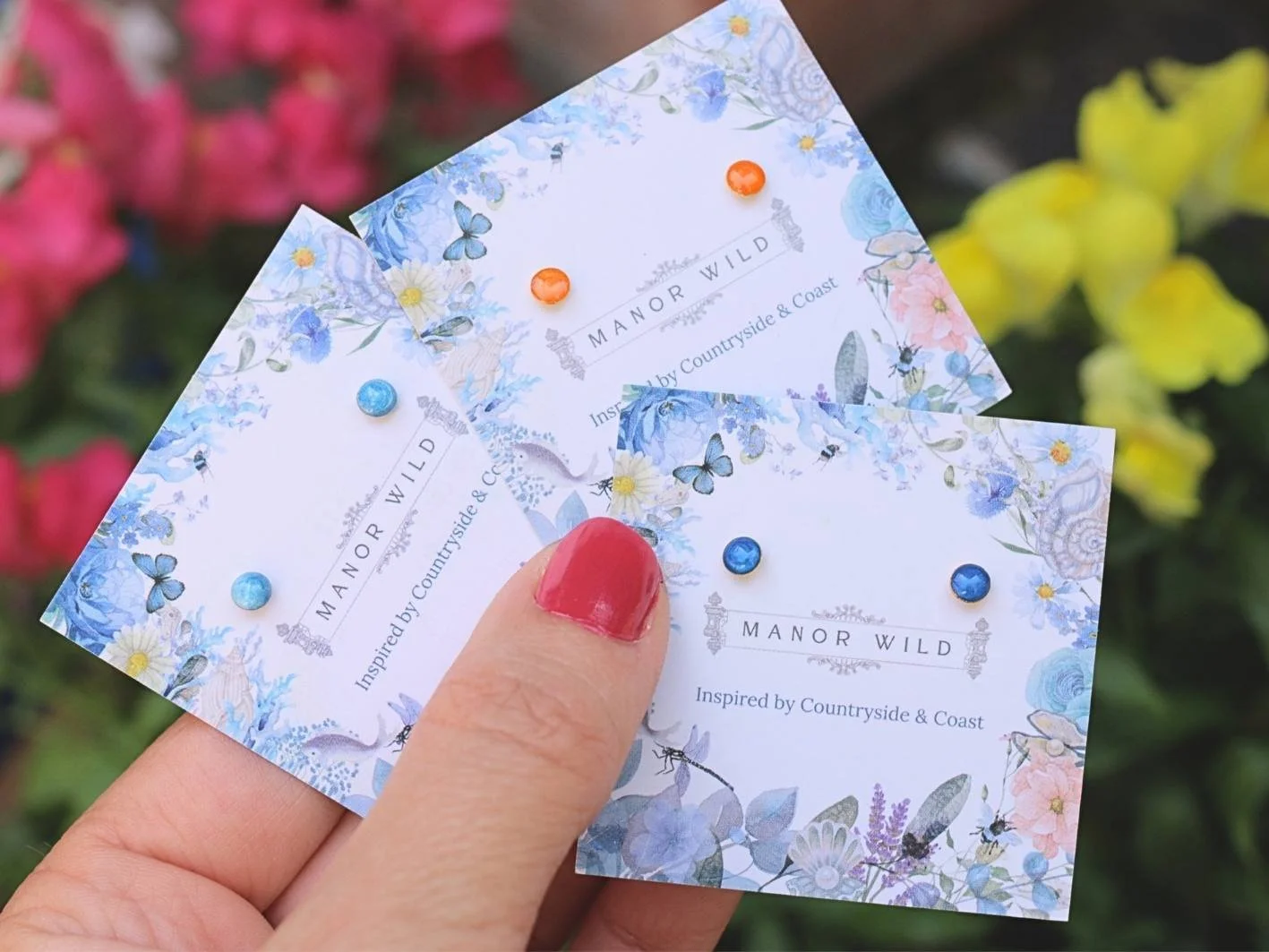 A hand with a red painted thumbnail holds three tickets for 'Manor Wild,' a brand inspired by countryside and coast, decorated with blue and pink floral patterns and small colored pins.
