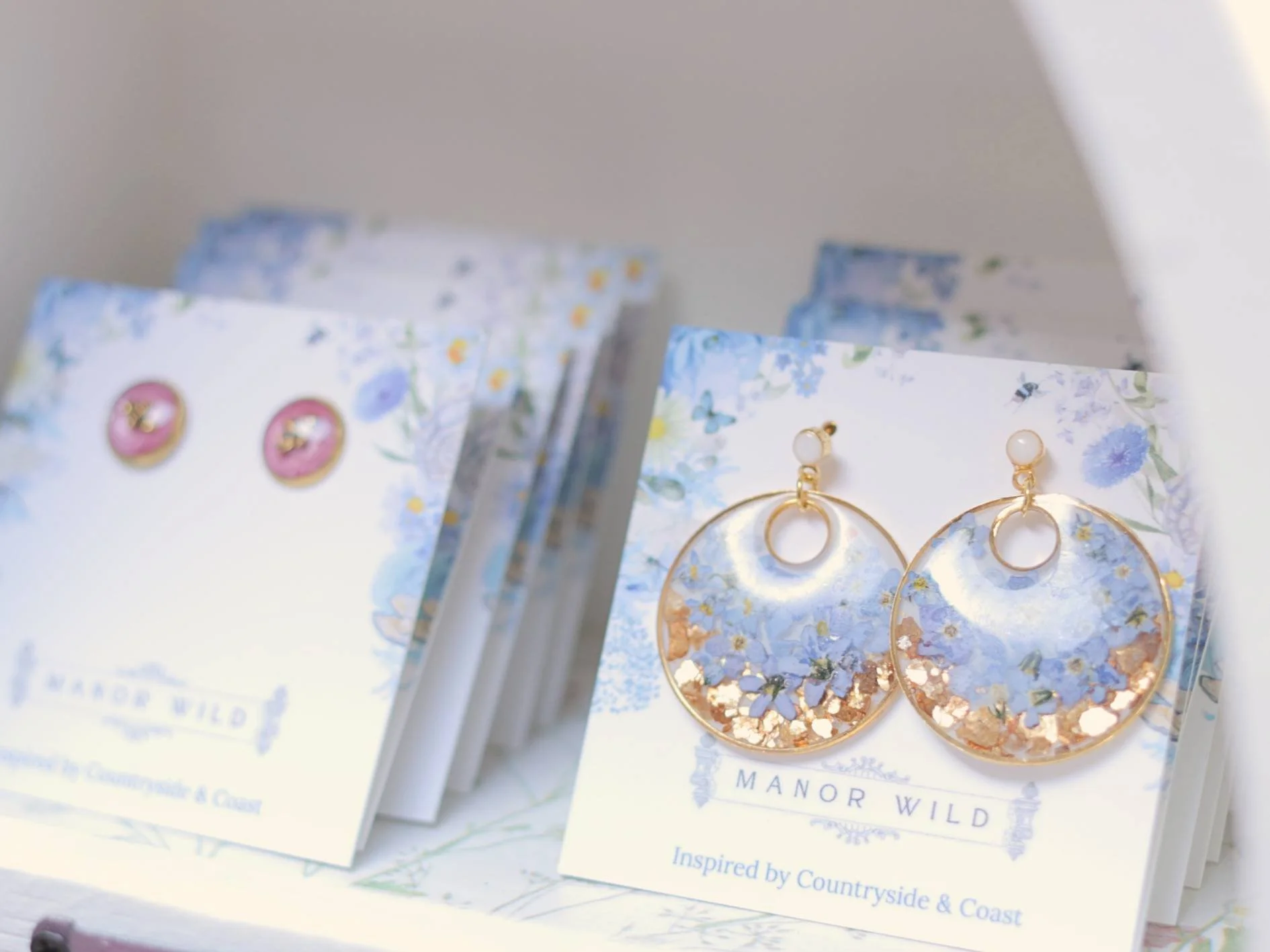 Pair of gold hoop earrings with white bead stud earrings on display, attached to cards with blue floral designs, in front of a partially blurred background showing more earring cards.