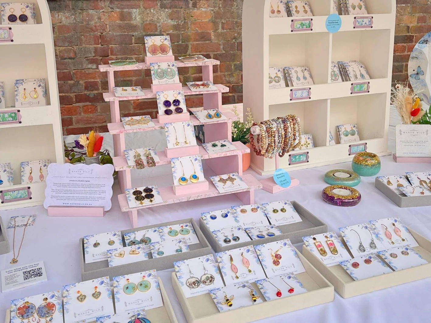 Display table of jewelry including earrings, necklaces, and bracelets arranged in trays and on shelves, against a brick wall background.