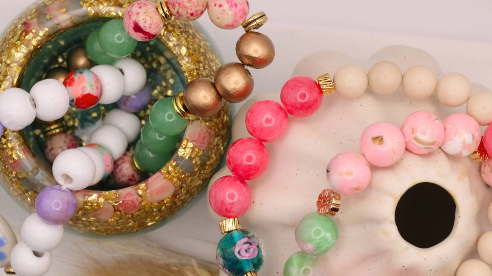 Colorful beaded necklaces and bracelets with pink, white, green, purple, and gold beads displayed on a white surface.