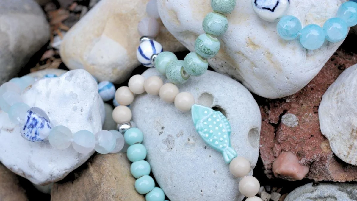Colorful beaded jewelry, including necklaces or bracelets with beads in shades of blue, green, and cream, resting on white and light-colored rocks or stones.
