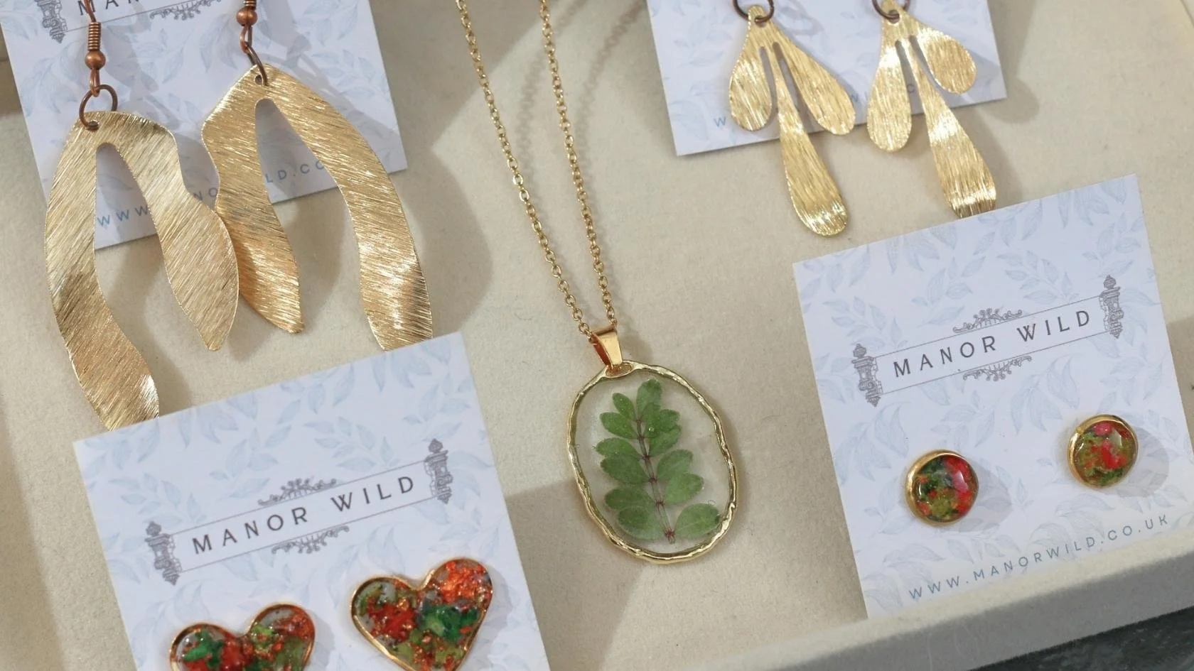 Gold leaf-shaped earrings, a leaf pendant necklace, and colorful heart-shaped earrings displayed on cards, with the brand name 'Manor Wild' visible.