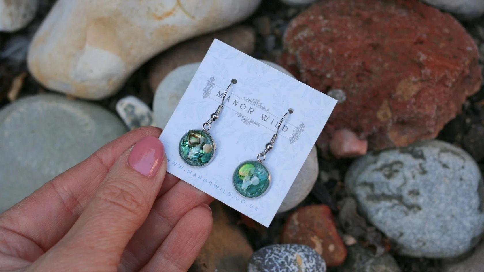 A hand holding a pair of earrings on a card from Manor Wild, with a rocky background.