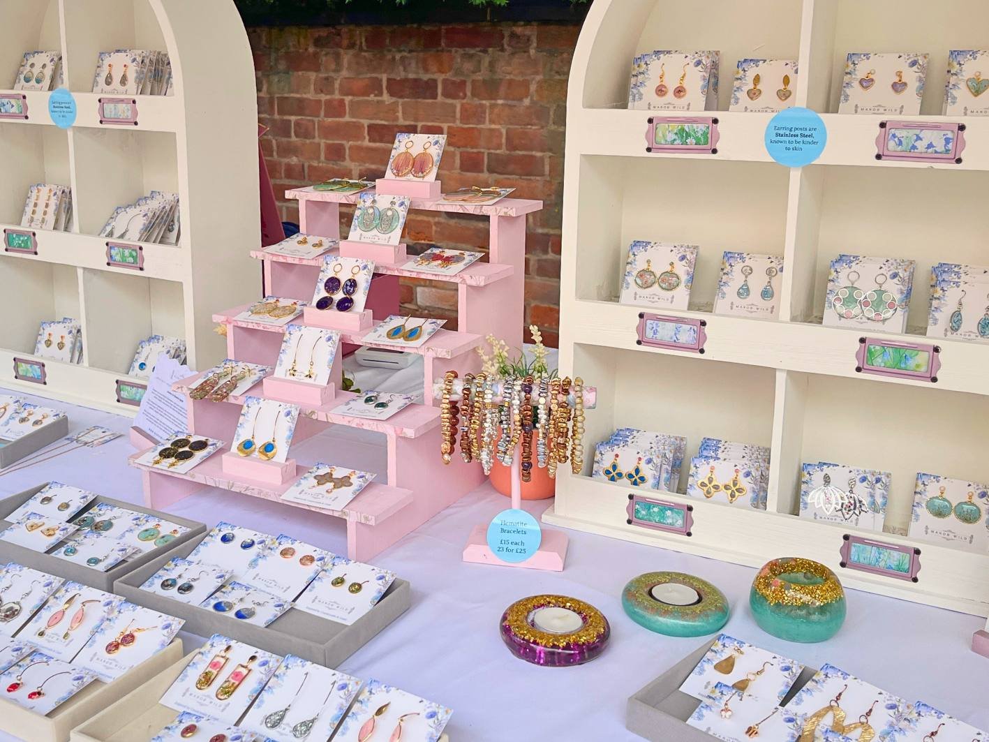Jewelry display at a market stall with earrings, necklaces, bracelets, and decorative candle holders on a white table with a brick wall in the background.