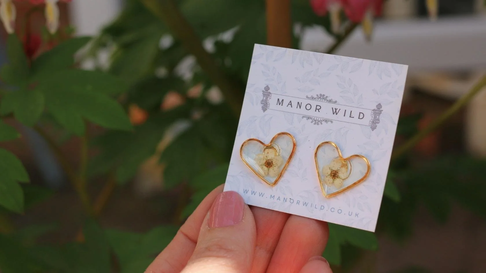 Hand holding a card with heart-shaped earrings containing small flowers, from Manor Wild