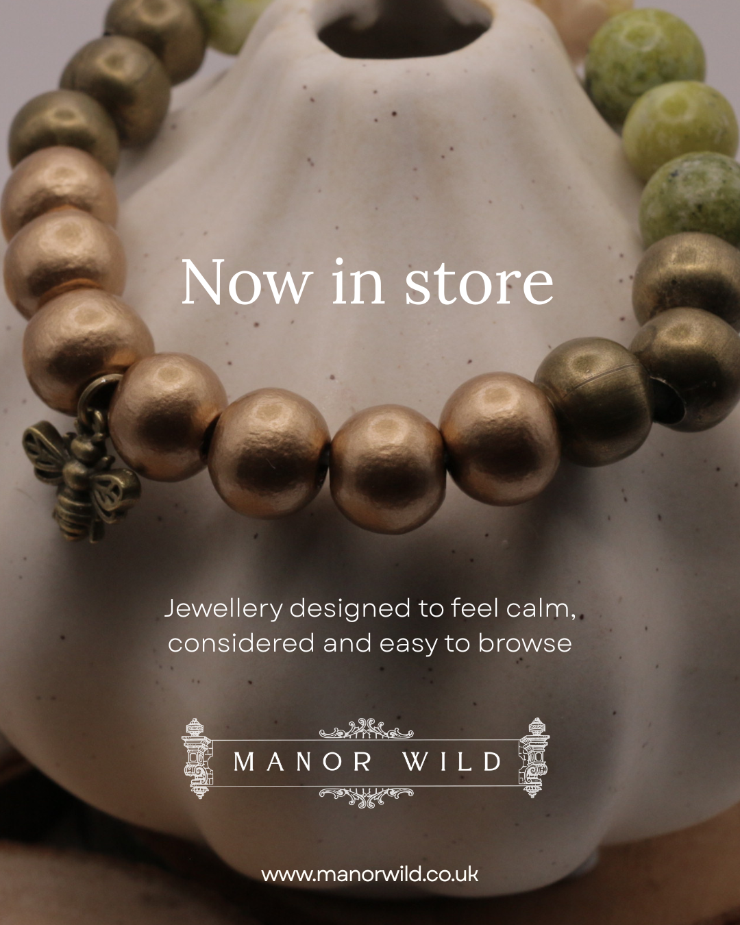Close-up photo of a beige ceramic vase with a beaded bracelet and a small bee charm, advertising Manor Wild jewellery now available in store.