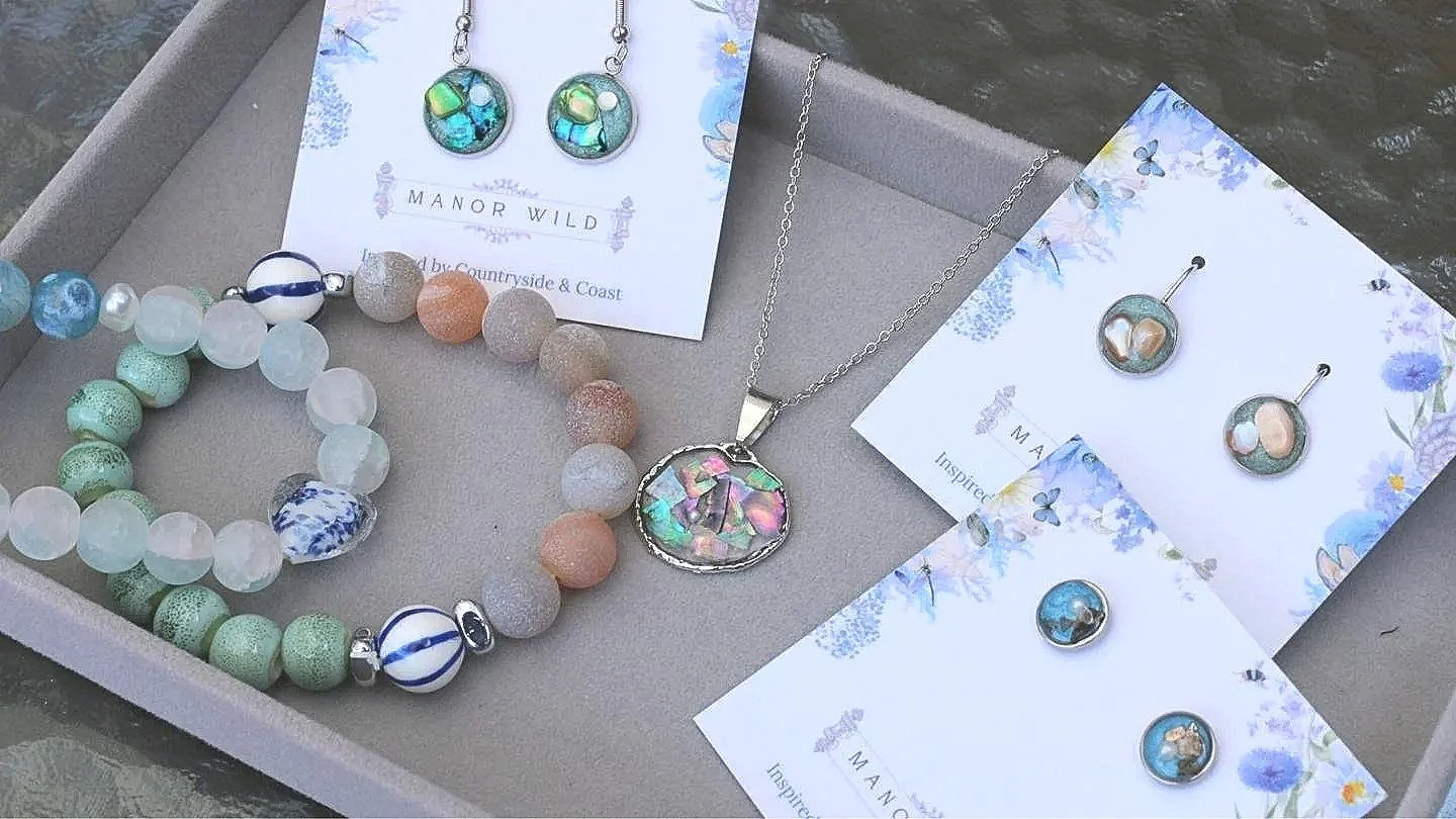 Jewelry set with earrings, necklace, and bracelet displayed in a gift box. The jewelry features colorful beads and floral designs, with cards labeled 'Manor Wild' and 'Inspired by Countryside & Coast'.