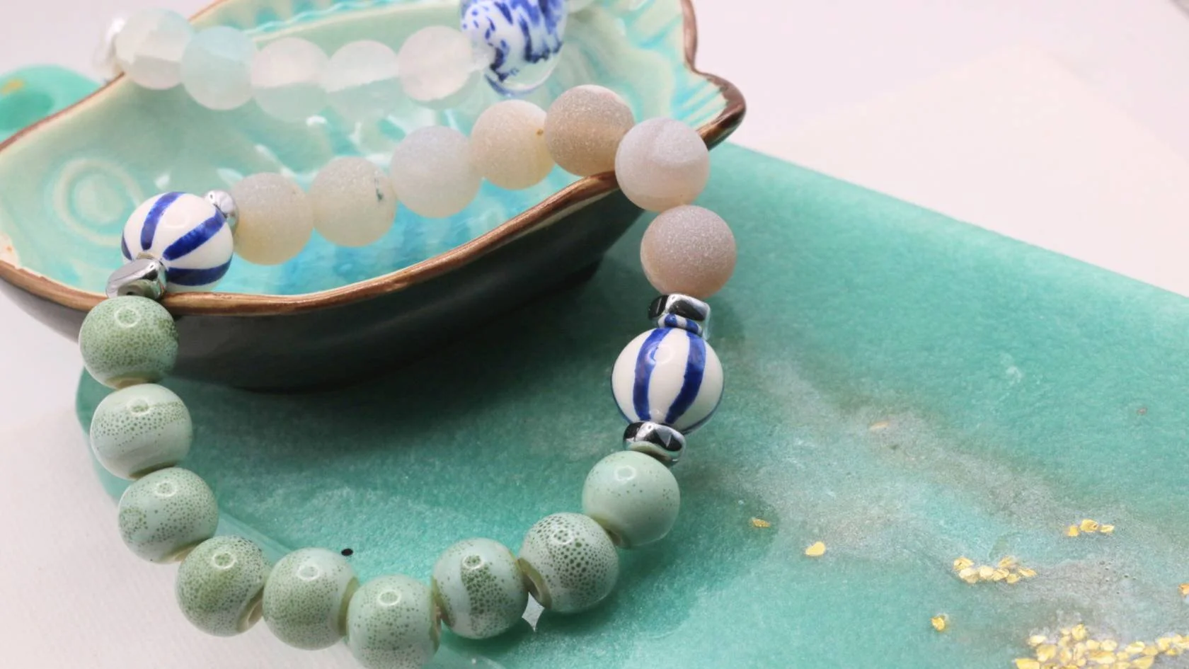 A necklace with spherical beads in white, green, and blue with striped patterns, resting on a green textured surface and next to a ceramic dish containing similar beads.