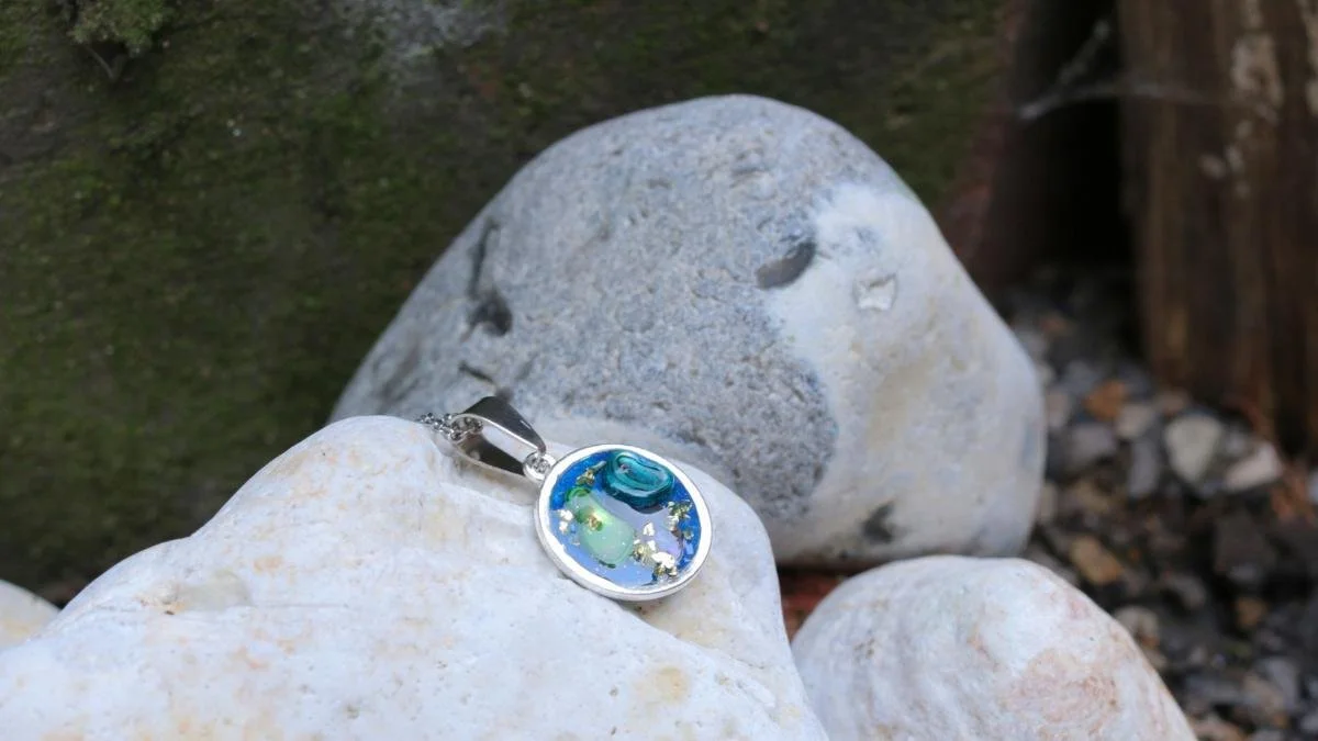 A small round pendant with a blue and green abstract design, resting on white rocks outdoors