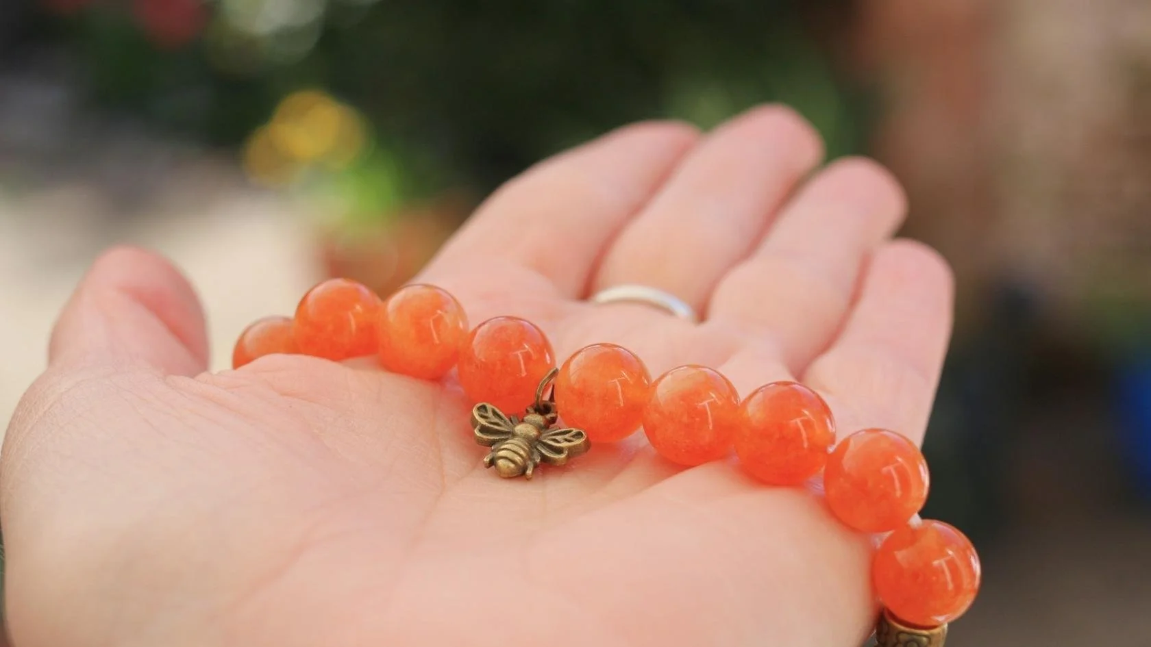 A person's open hand holding an orange beaded bracelet with a small bee charm. The background is blurry with hints of green, yellow, and blue.