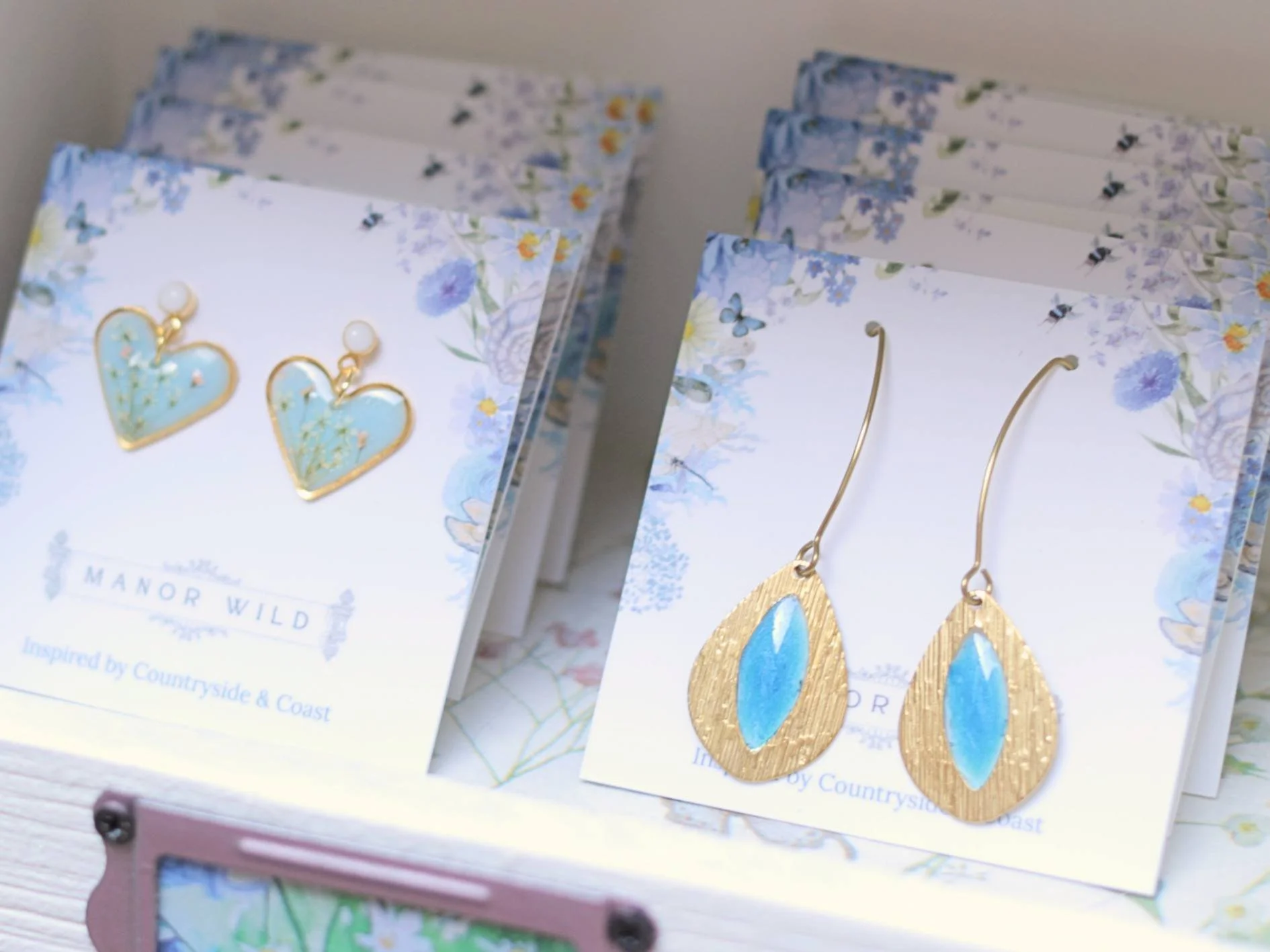 Close-up of jewelry earrings displayed on cards with floral designs, the cards are labeled "Manor Wild" and inspired by countryside and coast.