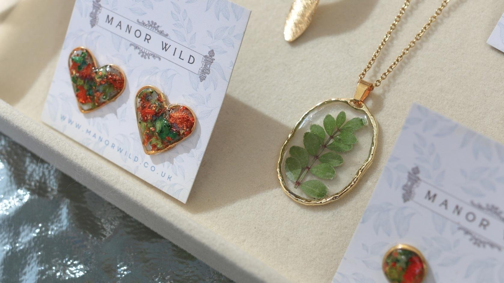 Gold and green leaf pendant necklace, colorful heart-shaped earrings with red, green, and gold accents, displayed on jewelry cards with the text "Manor Wild".