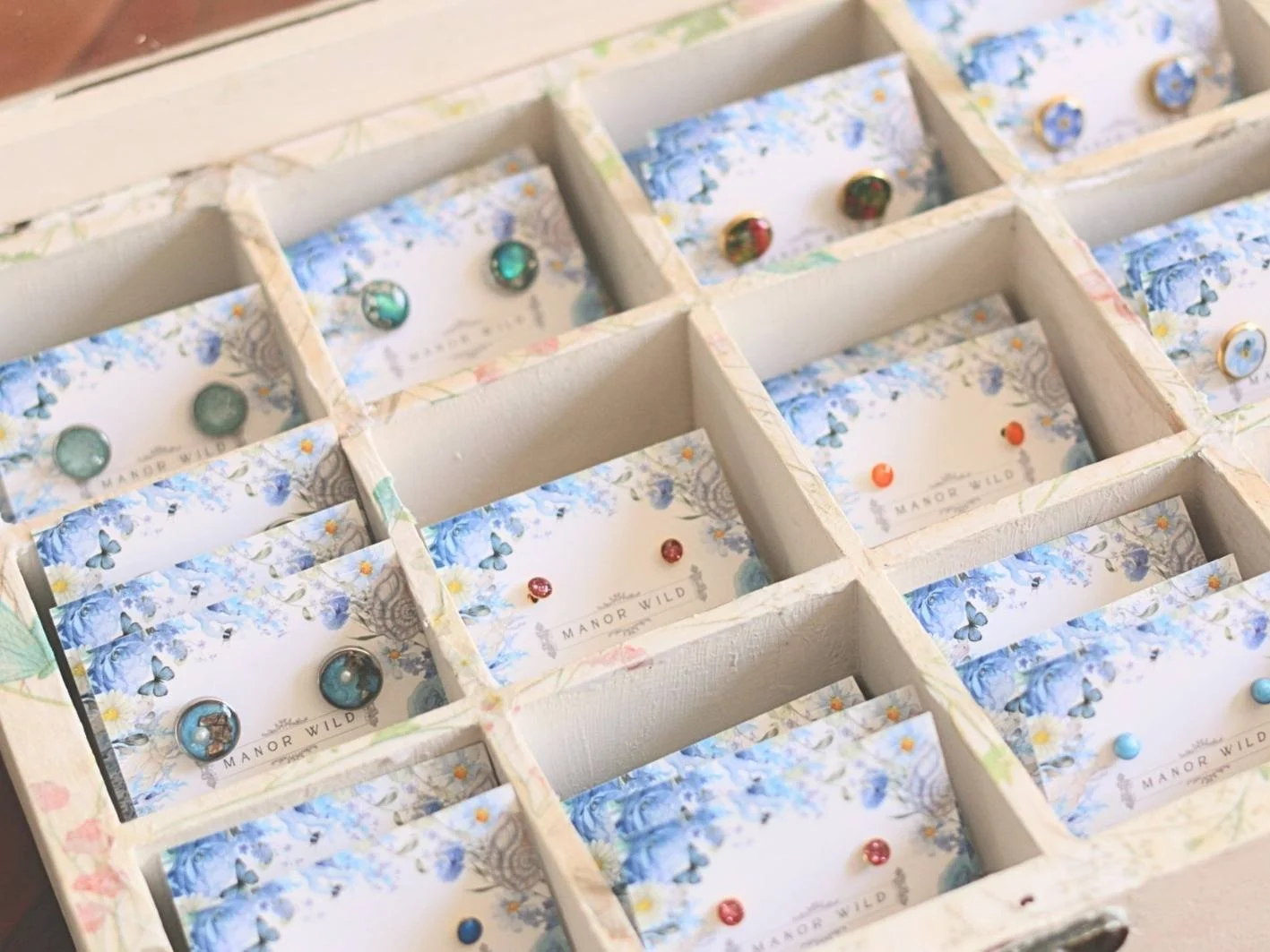 A white wooden box divided into compartments, each containing cards with blue floral and butterfly designs and the text 'Manor Wild,' decorated with colorful, assorted round beads.