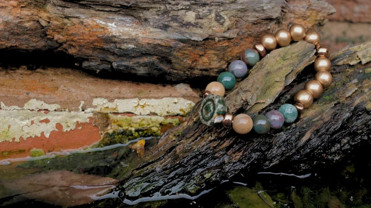 Beaded bracelet with various colored beads resting on a piece of wood near a brick wall and water.