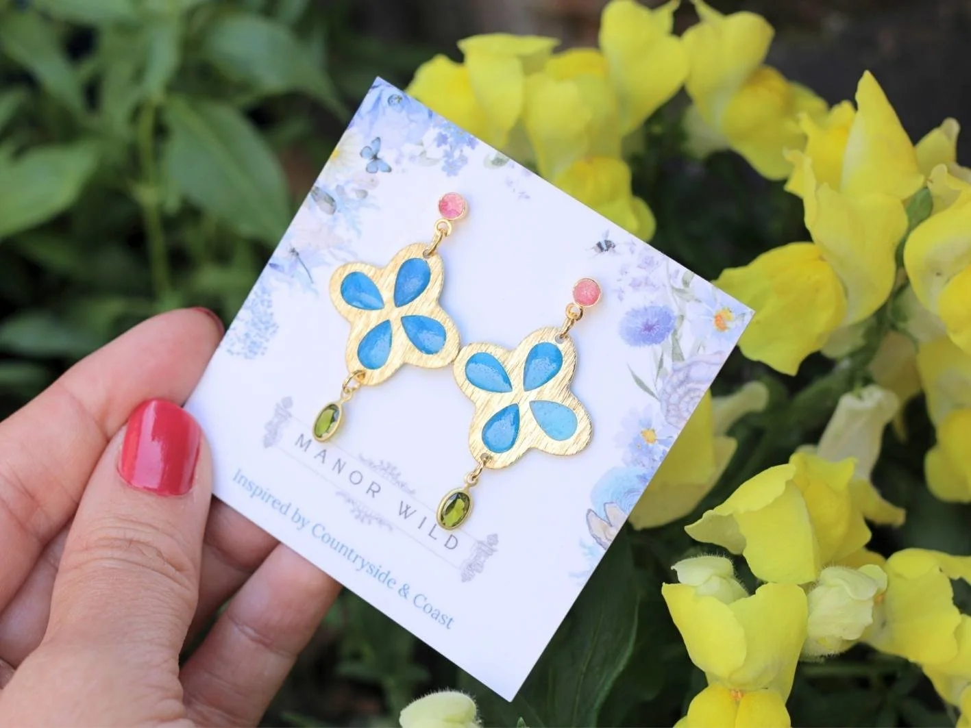 Colorful butterfly earrings with blue, pink, and green stones on a white card, held by a person with pink nail polish, outdoors near yellow flowers.