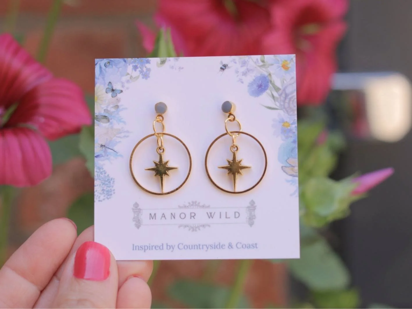 Gold earrings with a star-shaped charm inside a circular hoop, displayed on a floral card labeled 'Manor Wild' with the tagline 'Inspired by Countryside & Coast', held by a person with pink painted nails, with pink flowers in the background.