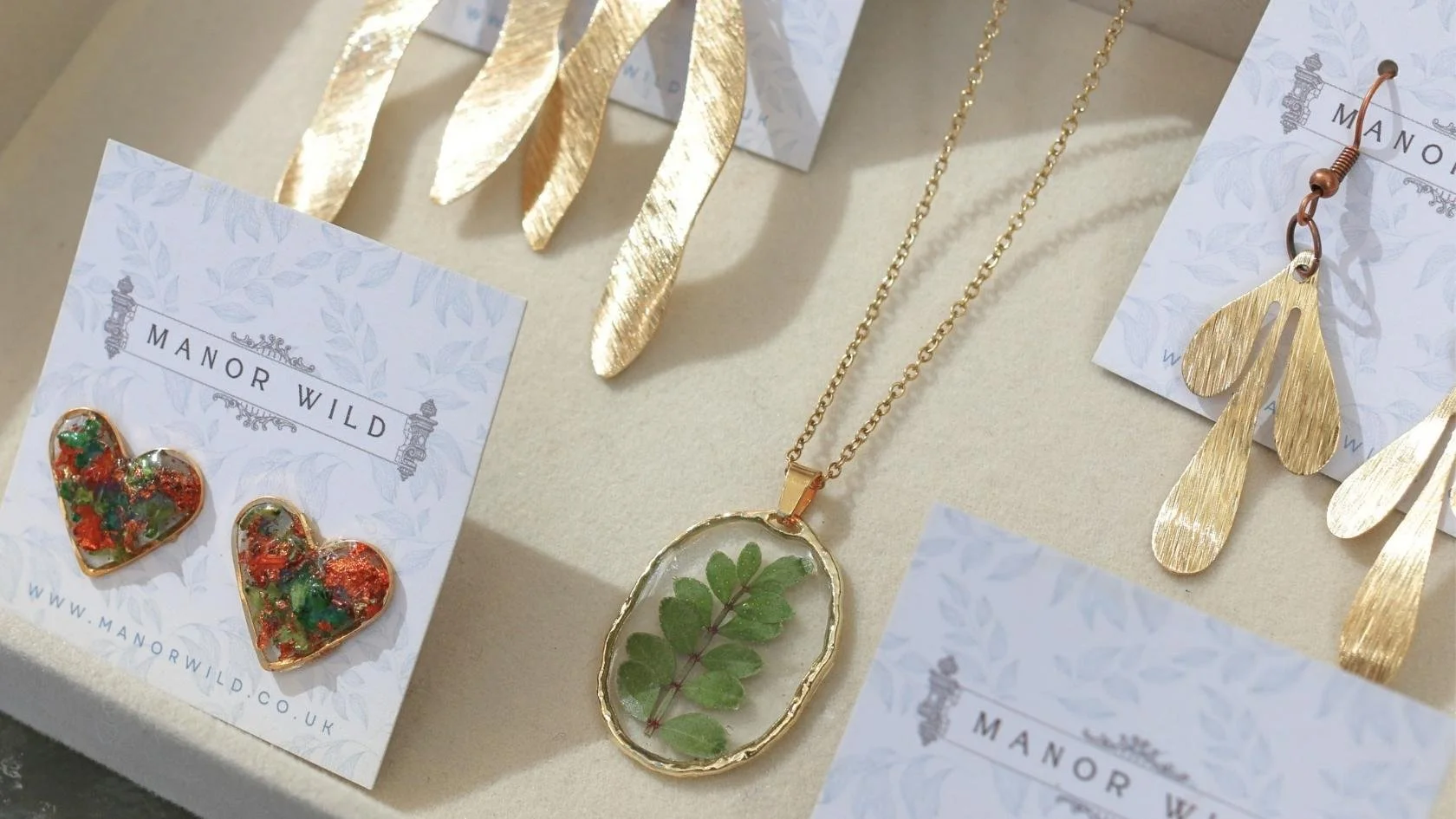 Gold jewelry including earrings and necklaces with leaf and floral designs, displayed on white cards with "Manor Wild" branding.