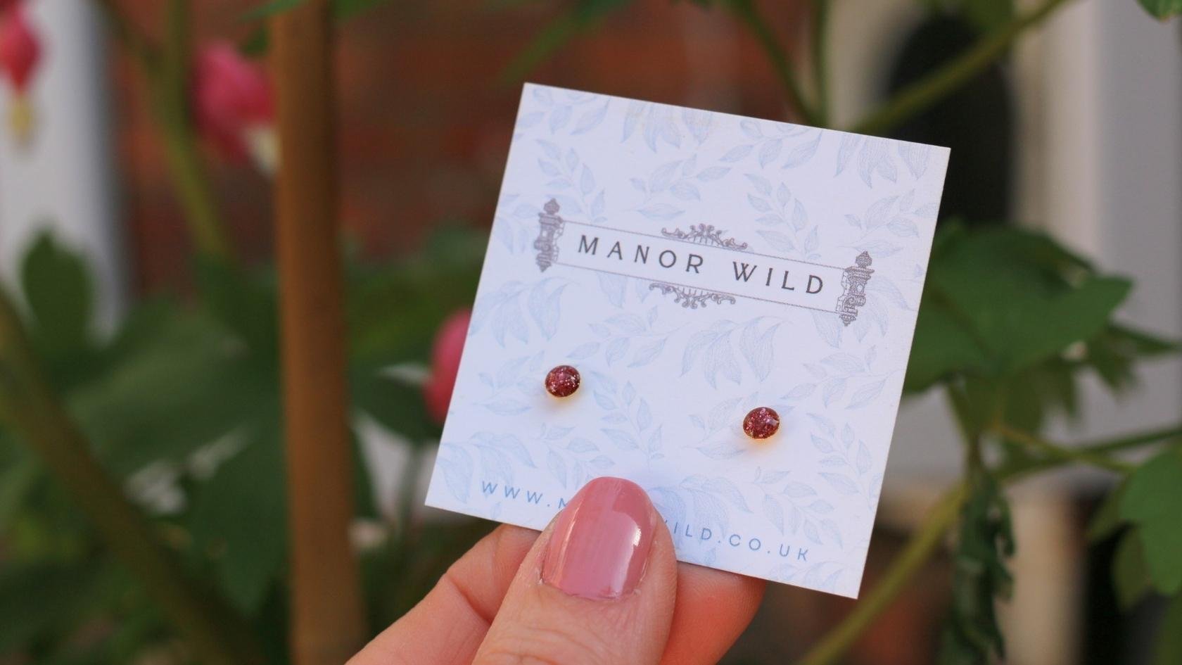 A hand holding a white card with the text 'Manor Wild' and a website URL, displaying a pair of small red earrings in front of green plants.
