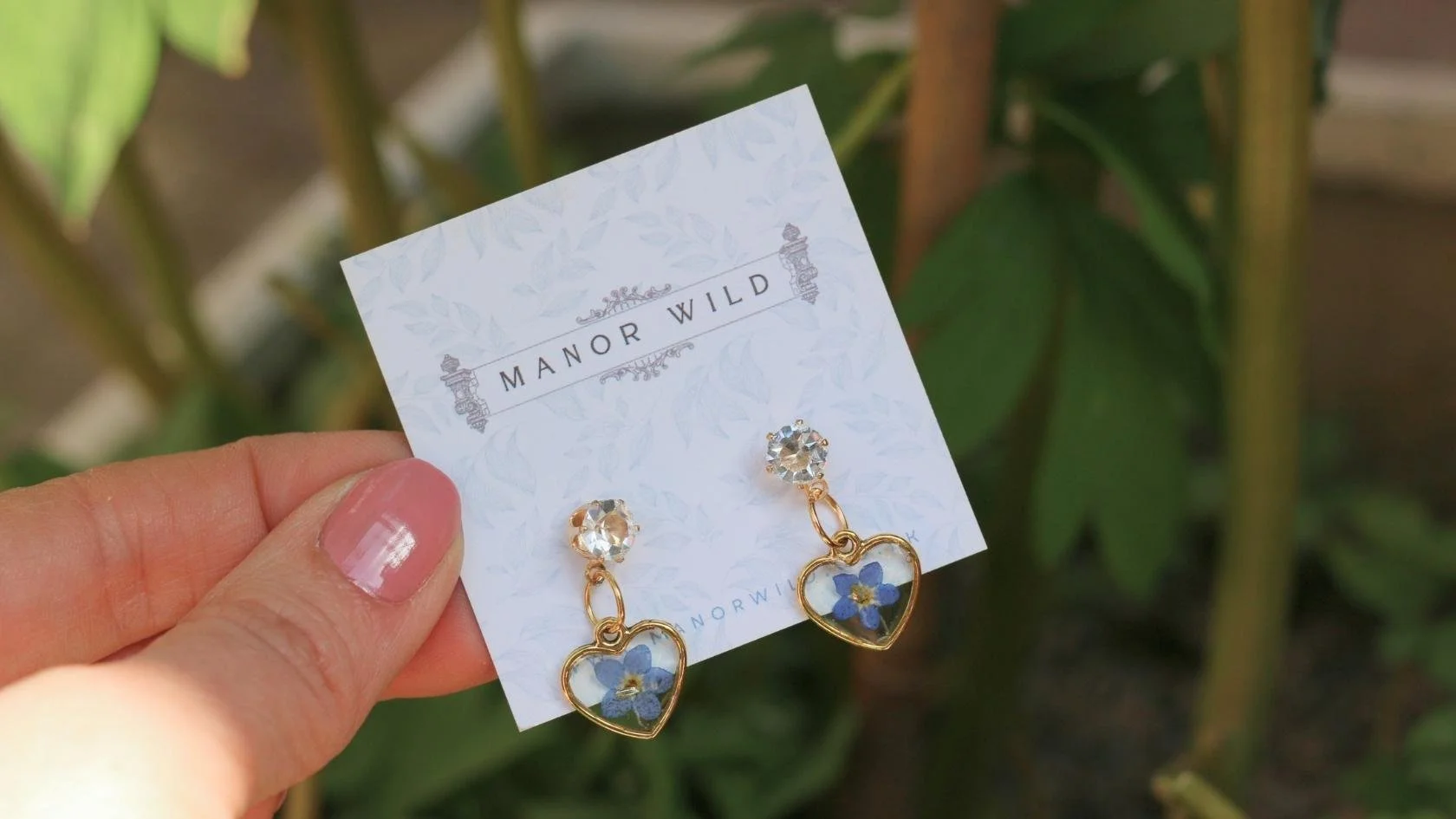 A hand holding a pair of gold earrings with clear stones and heart-shaped pendants containing blue flowers on a white card with 'Manor Wild' written on it, outdoors with green foliage in the background.