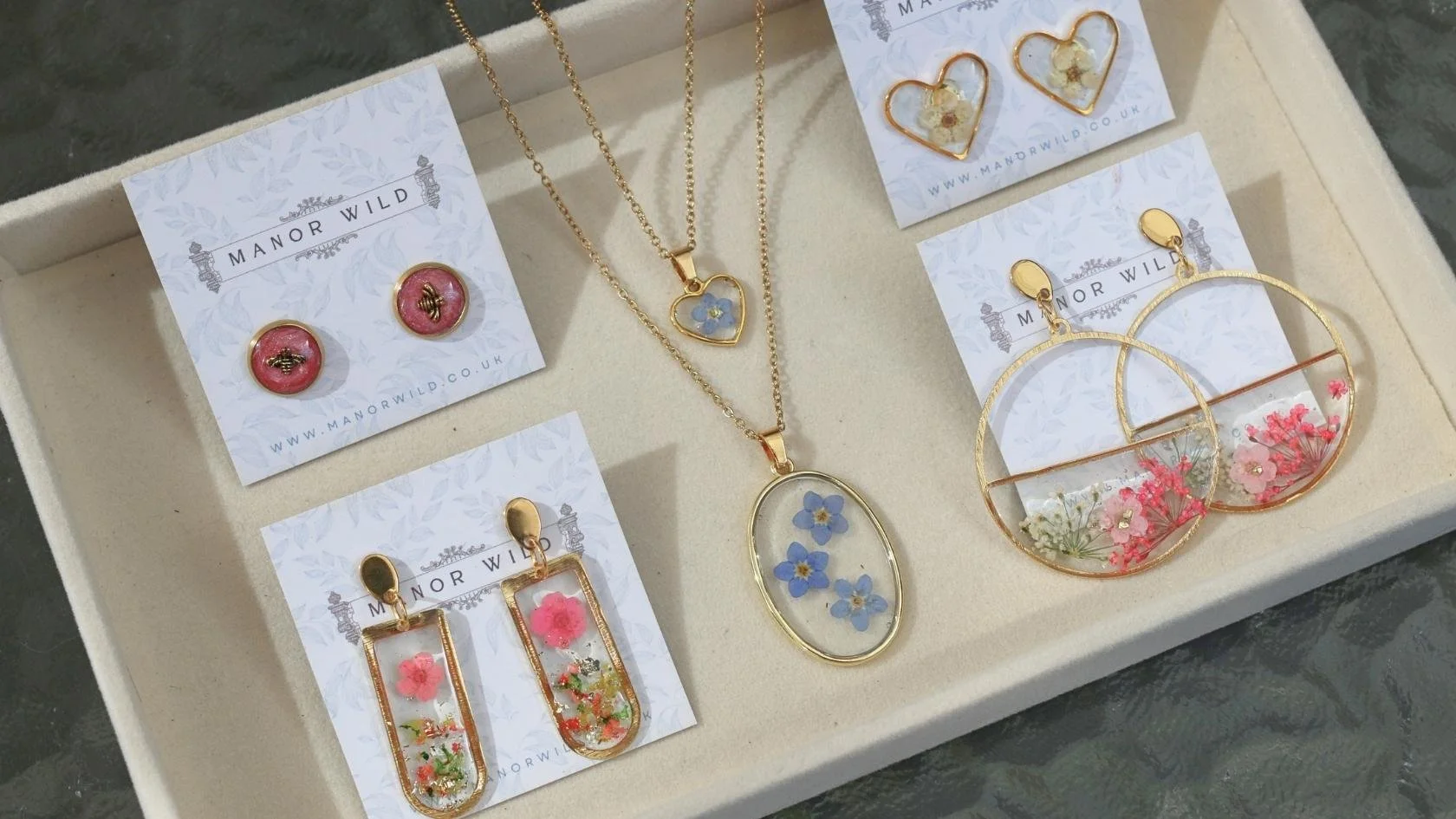 A jewelry display tray with floral jewelry including earrings and necklaces containing pressed flowers set in gold-tone metal, from Manor Wild.