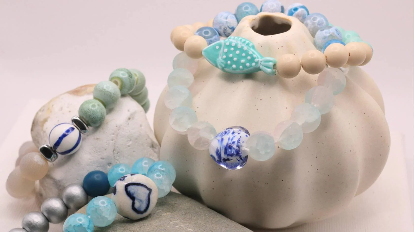 Several beaded necklaces and bracelets in shades of blue, white, and silver, displayed on smooth stones and a cream-colored ceramic vessel.